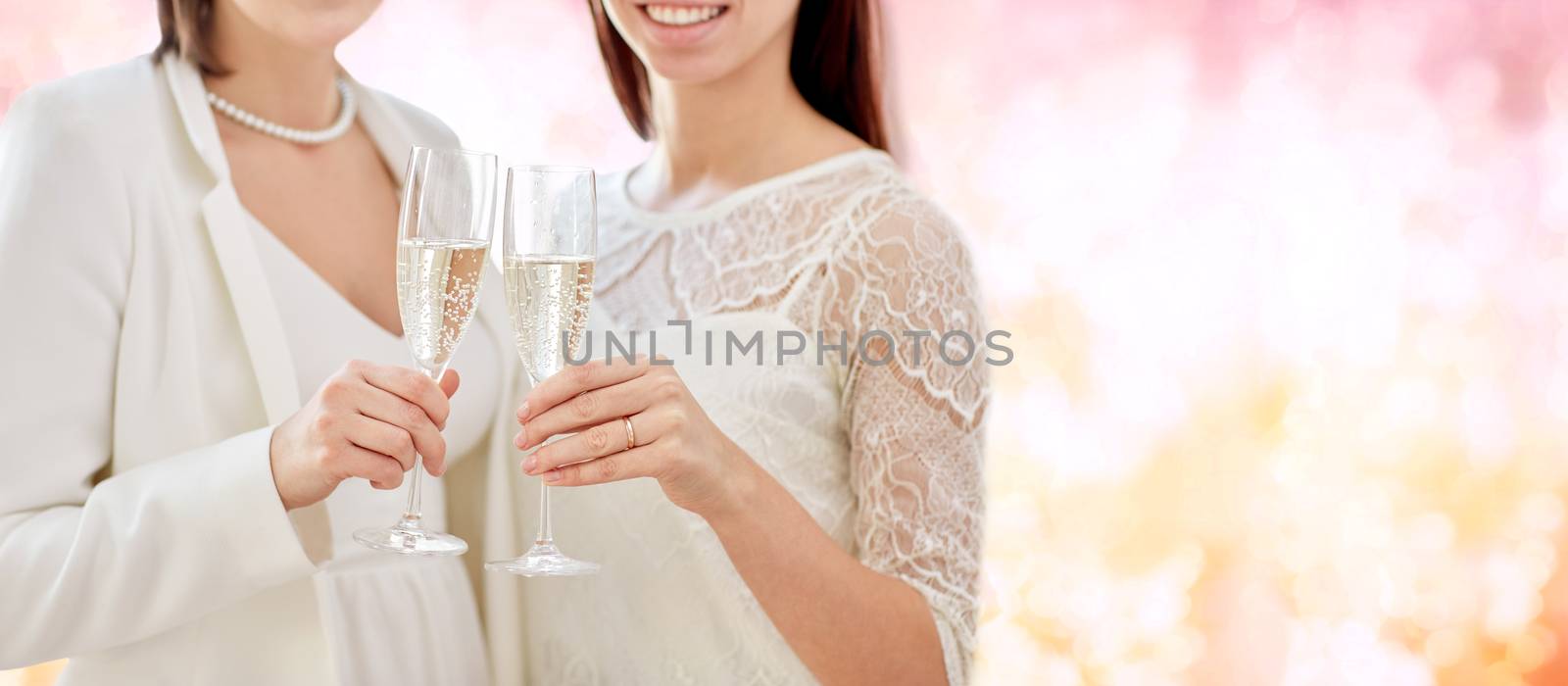 people, homosexuality, same-sex marriage, celebration and love concept - close up of happy married lesbian couple holding and clinking champagne glasses over pink holidays lights background