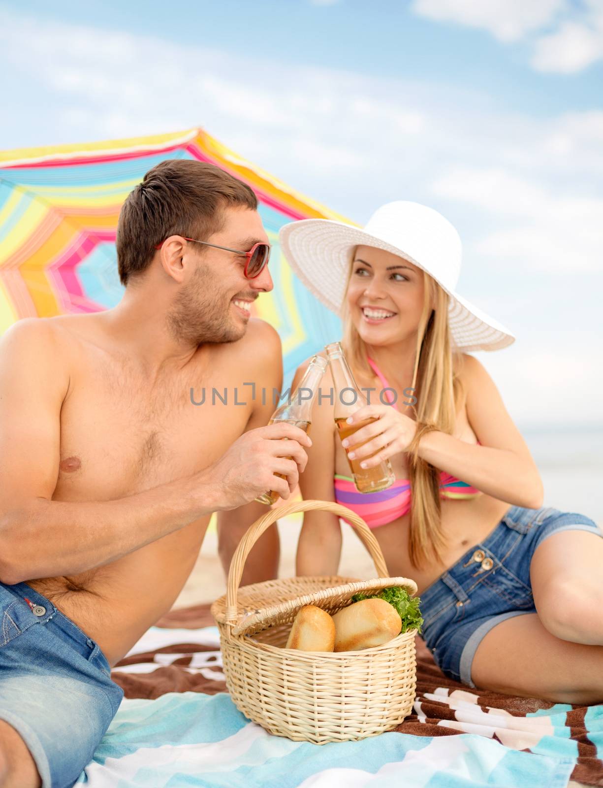summer, holidays, vacation and happy people concept - happy couple with bottle drinks having picnic and sunbathing on beach under colorful parasol
