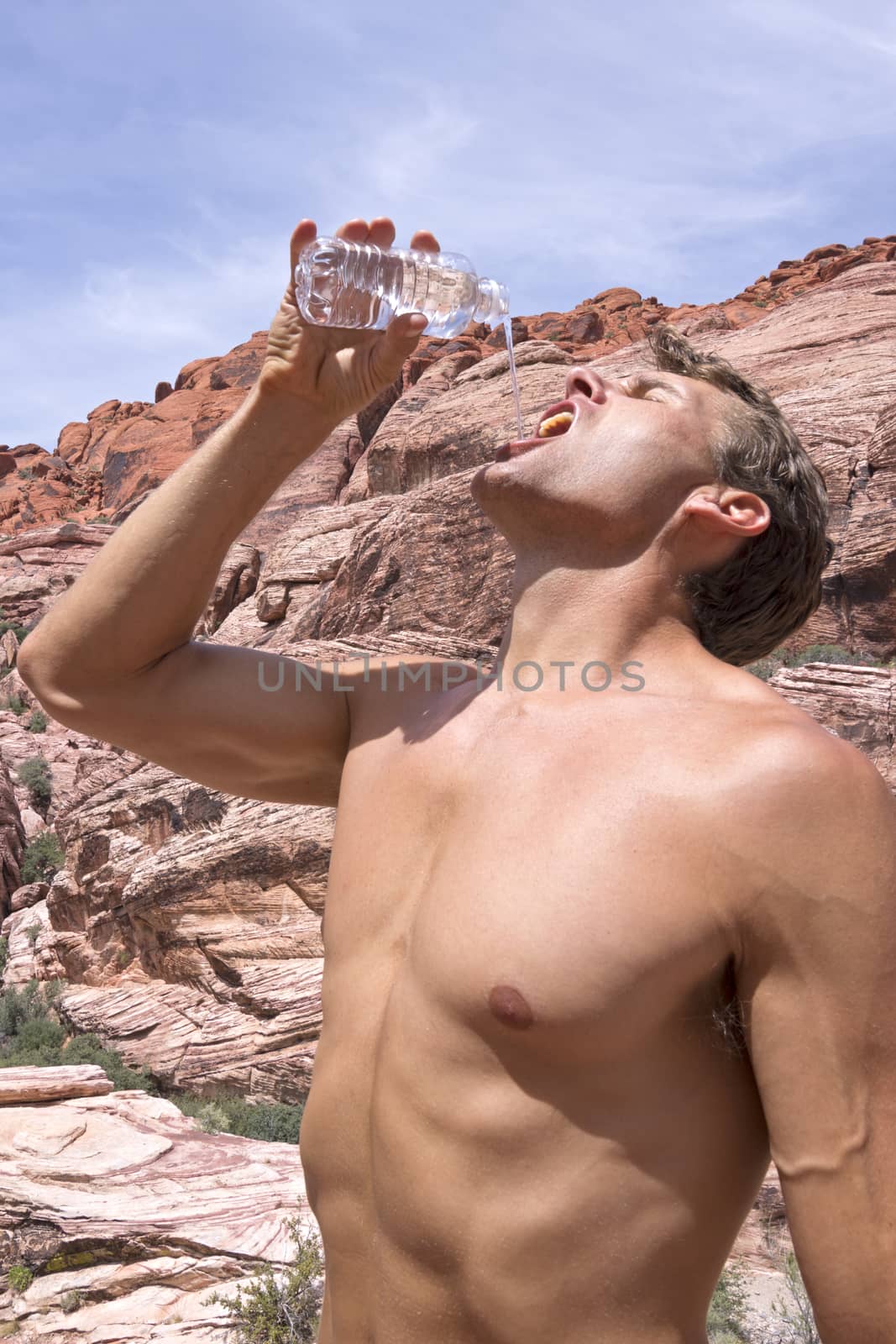 Muscular shirtless Caucasian man tilts back head and pours drinking water into mouth from plastic bottle while hiking in Red Rock Canyon desert in Las Vegas on hot sunny day