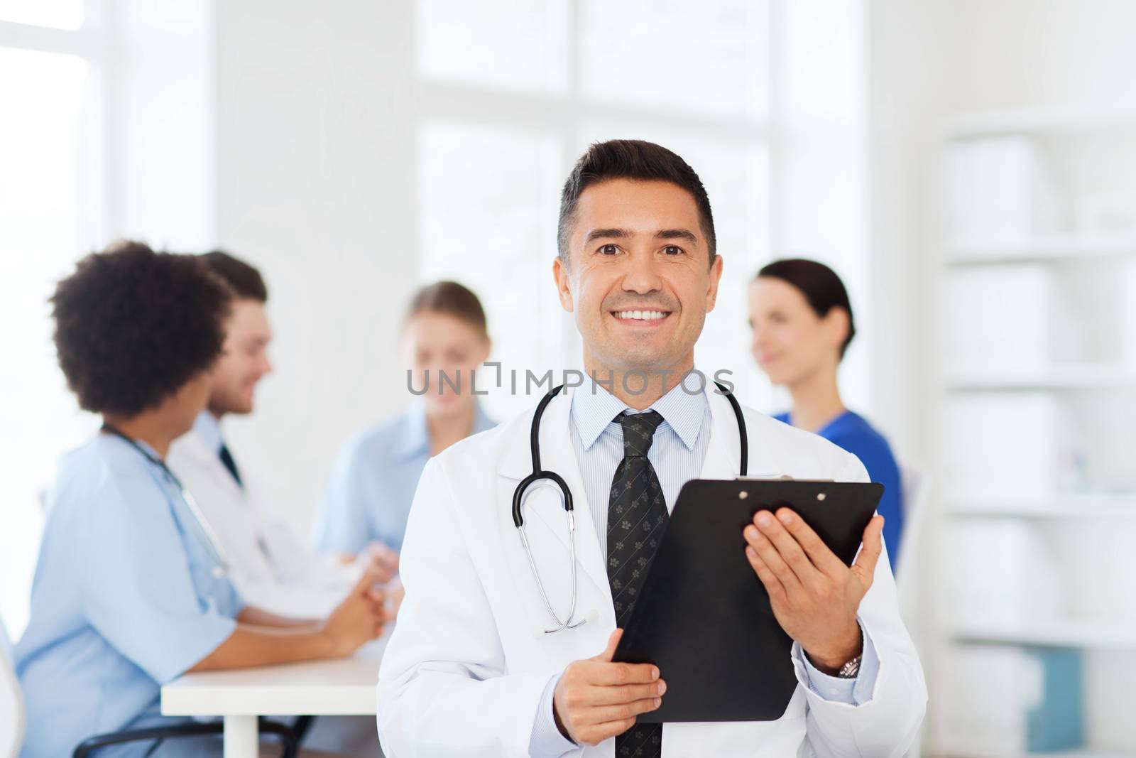 clinic, profession, people and medicine concept - happy male doctor with clipboard over group of medics meeting at hospital