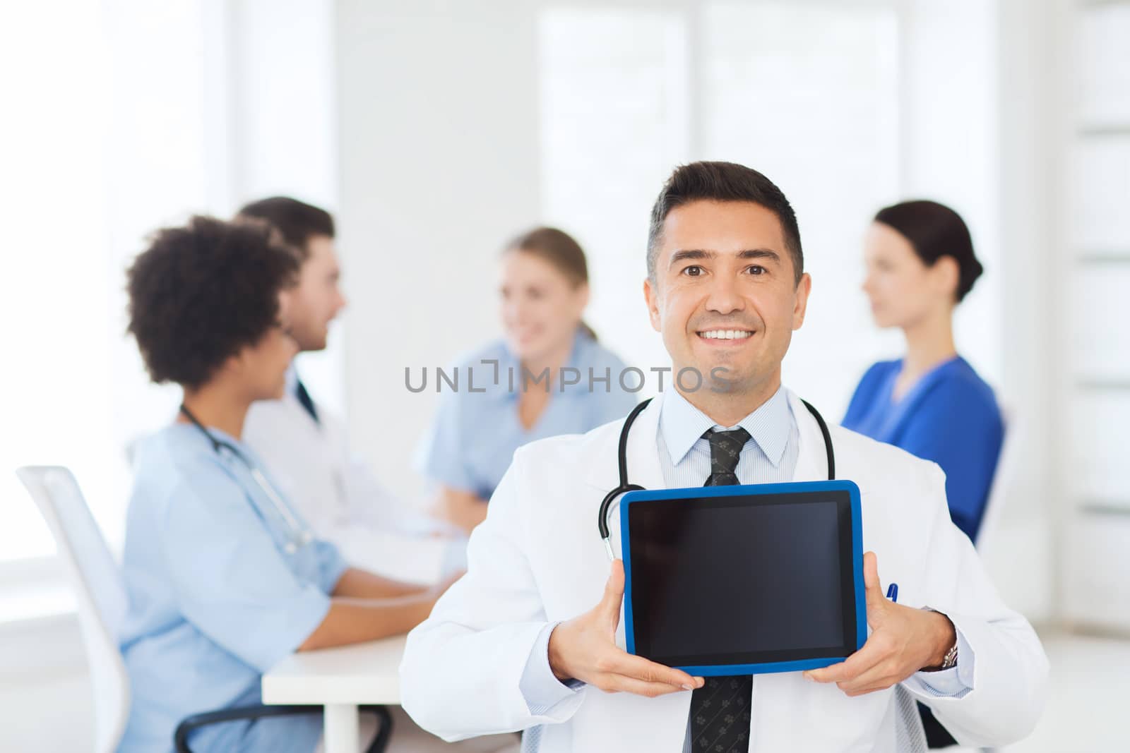 clinic, profession, people and medicine concept - happy male doctor showing tablet pc computer blank screen over group of medics meeting at hospital