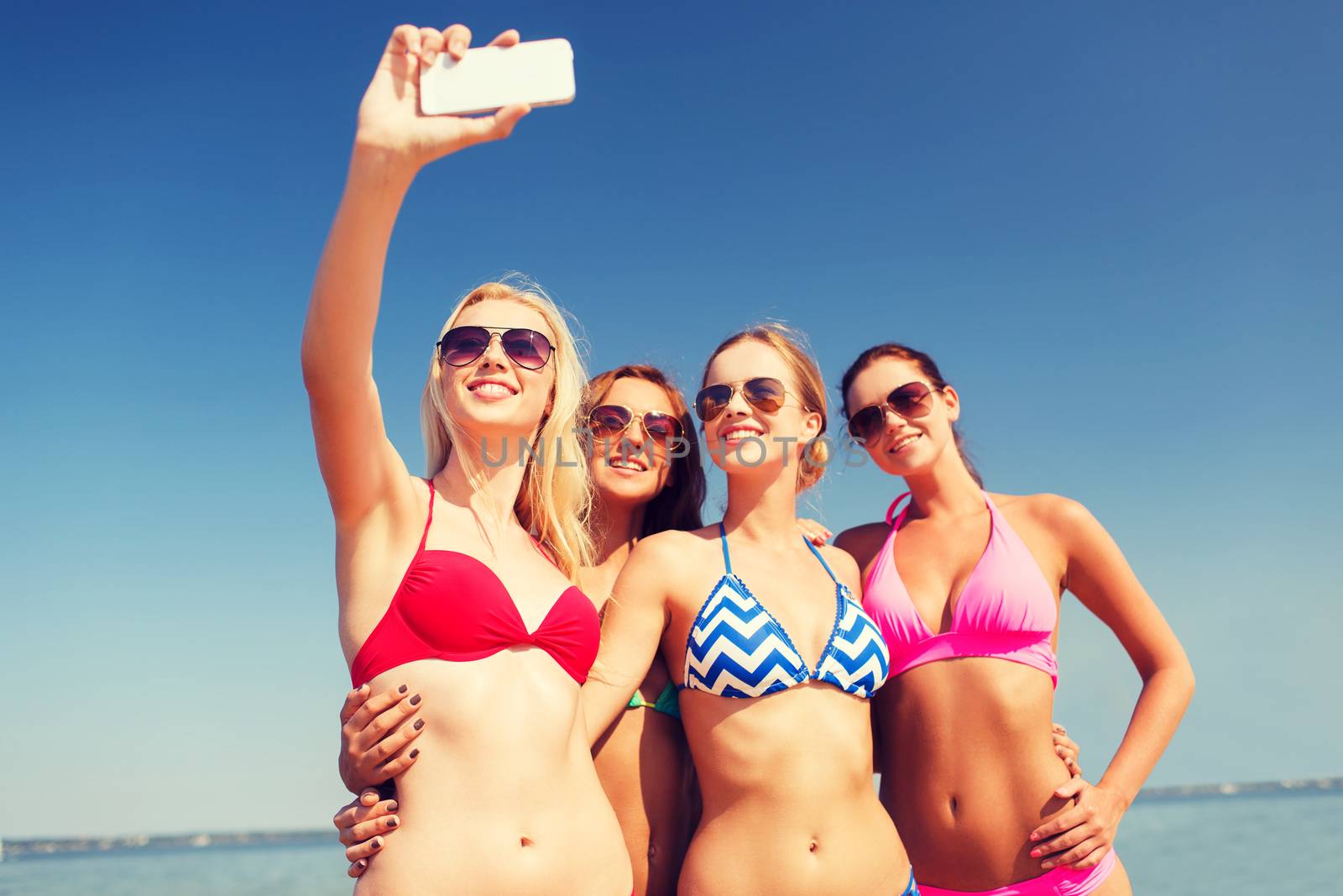 summer vacation, holidays, travel, technology and people concept- group of smiling young women on beach making selfie with smartphone over blue sky background