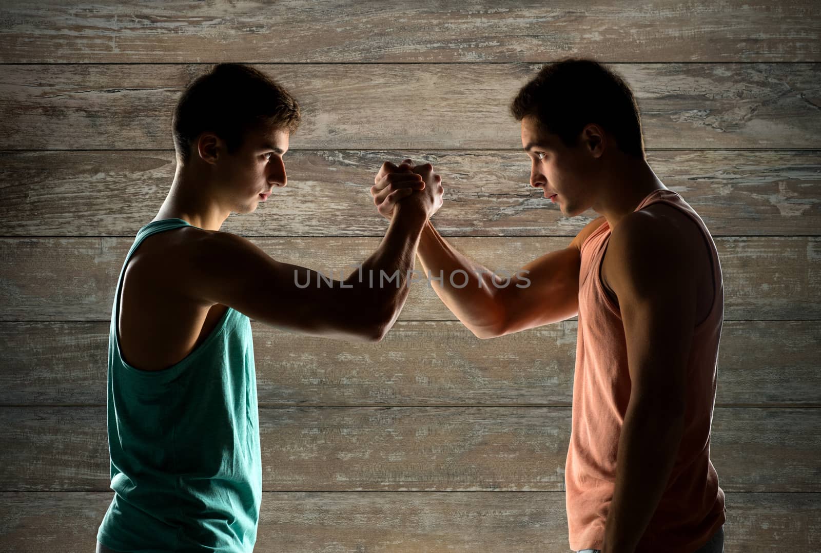 sport, competition, strength and people concept - two young men arm wrestling over wooden wall background