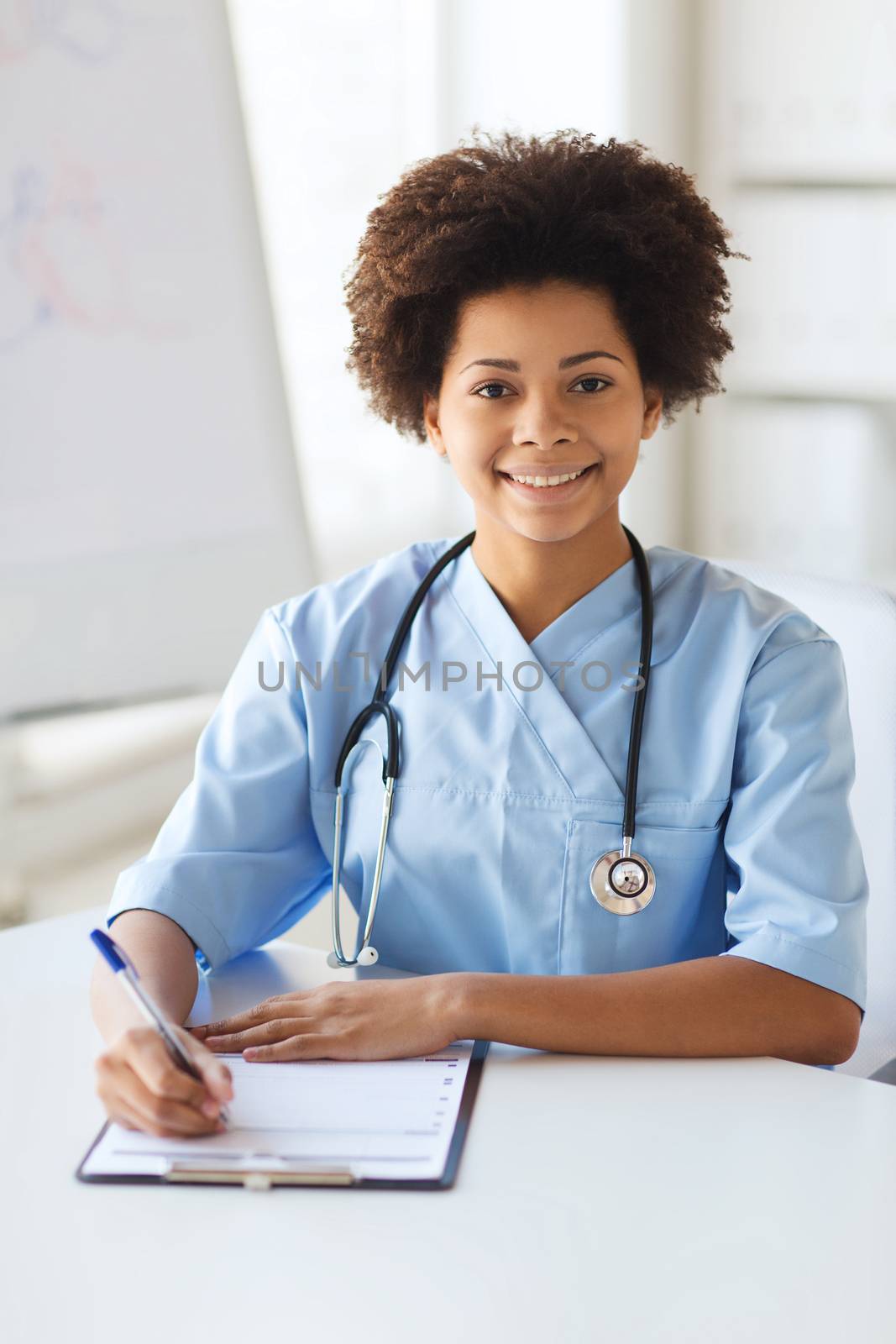 medicine, people and healthcare concept - happy female african american doctor or nurse writing medical report to clipboard at hospital