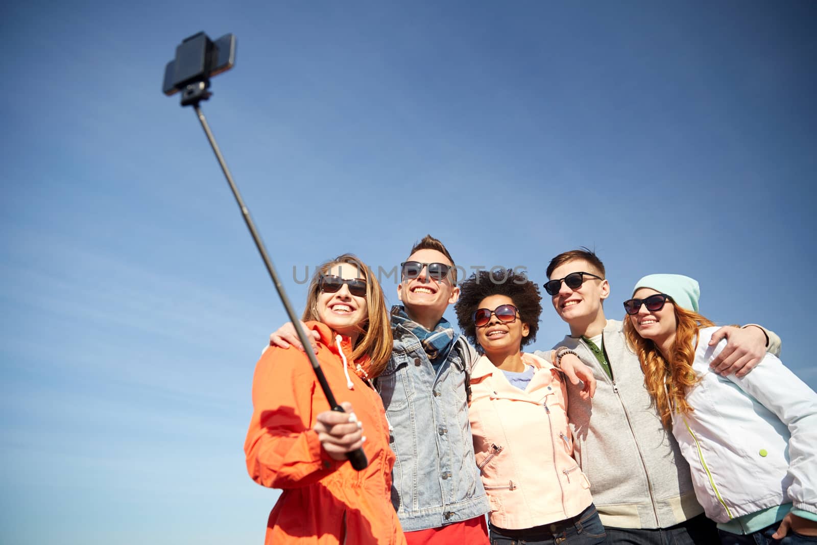 tourism, travel, people, leisure and technology concept - group of smiling teenage friends taking selfie with smartphone and monopod outdoors