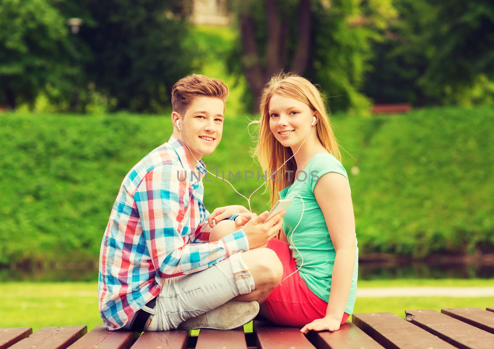summer, vacation, holidays, technology and friendship concept - smiling couple with smartphone and earphones sitting on bench in park