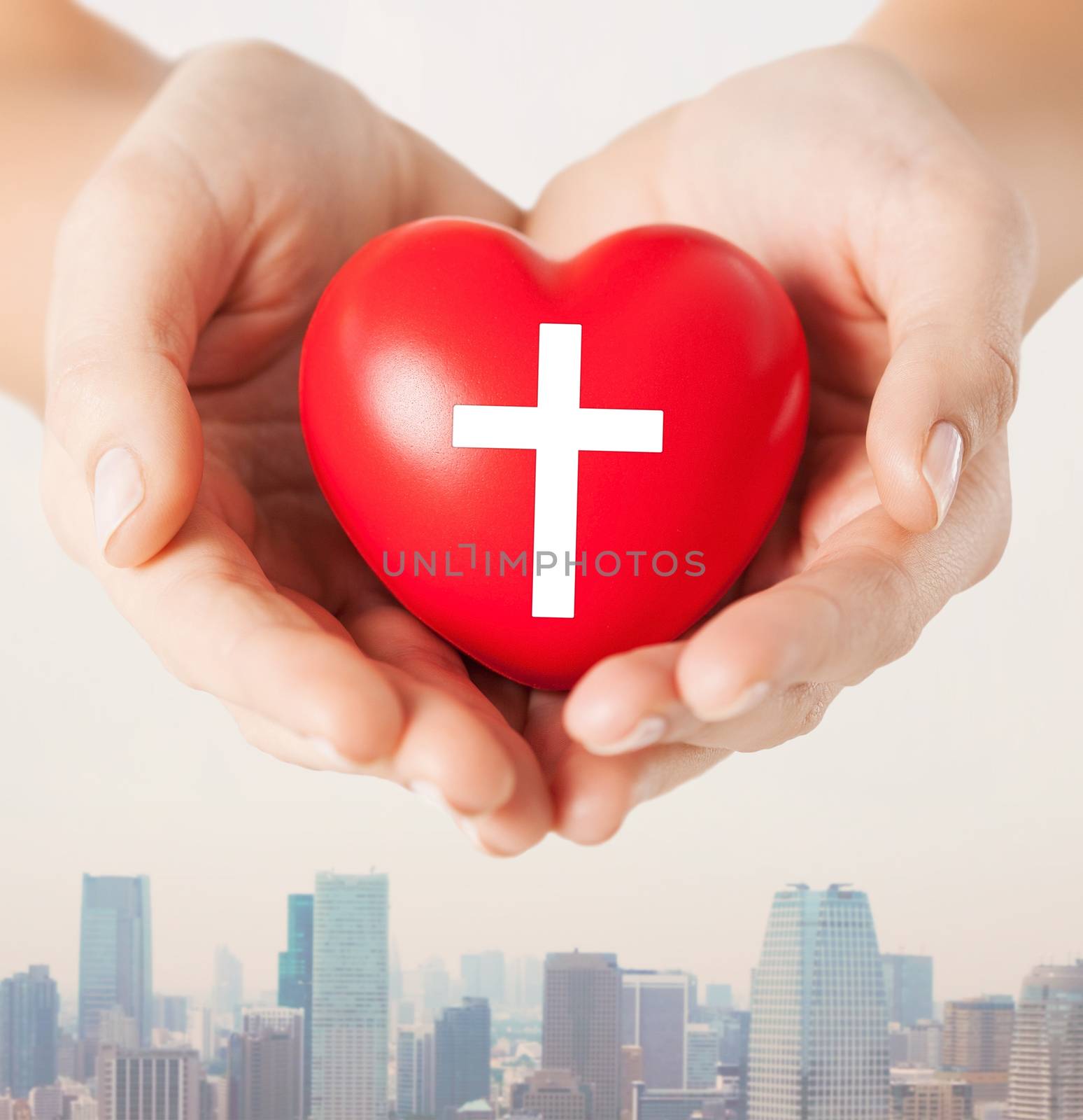 religion, christianity and charity concept - close up of female hands holding red heart with christian cross symbol over city skyscrapers background