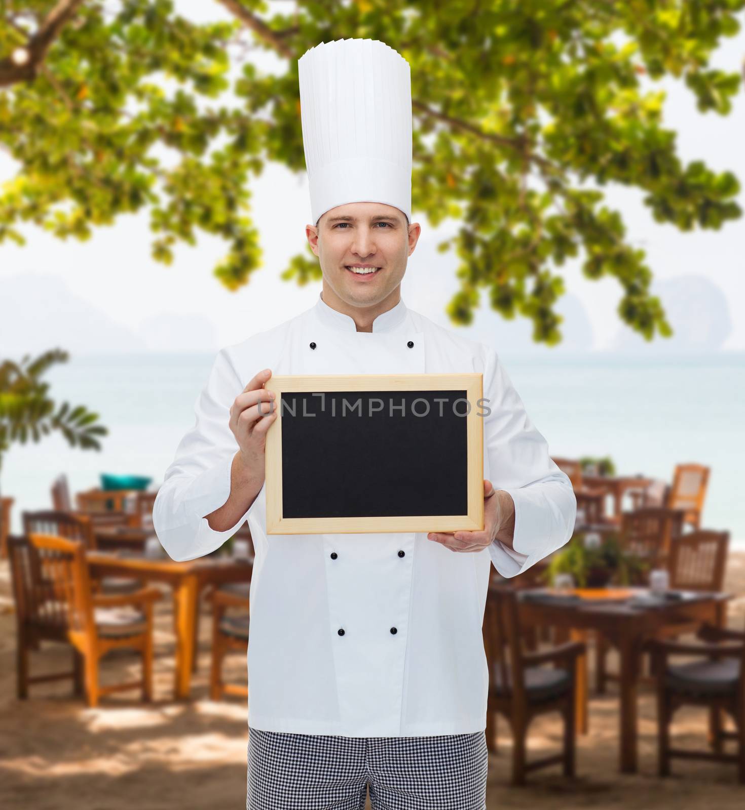 cooking, profession, advertisement and people concept - happy male chef cook showing and holding blank menu board over restaurant lounge on beach
