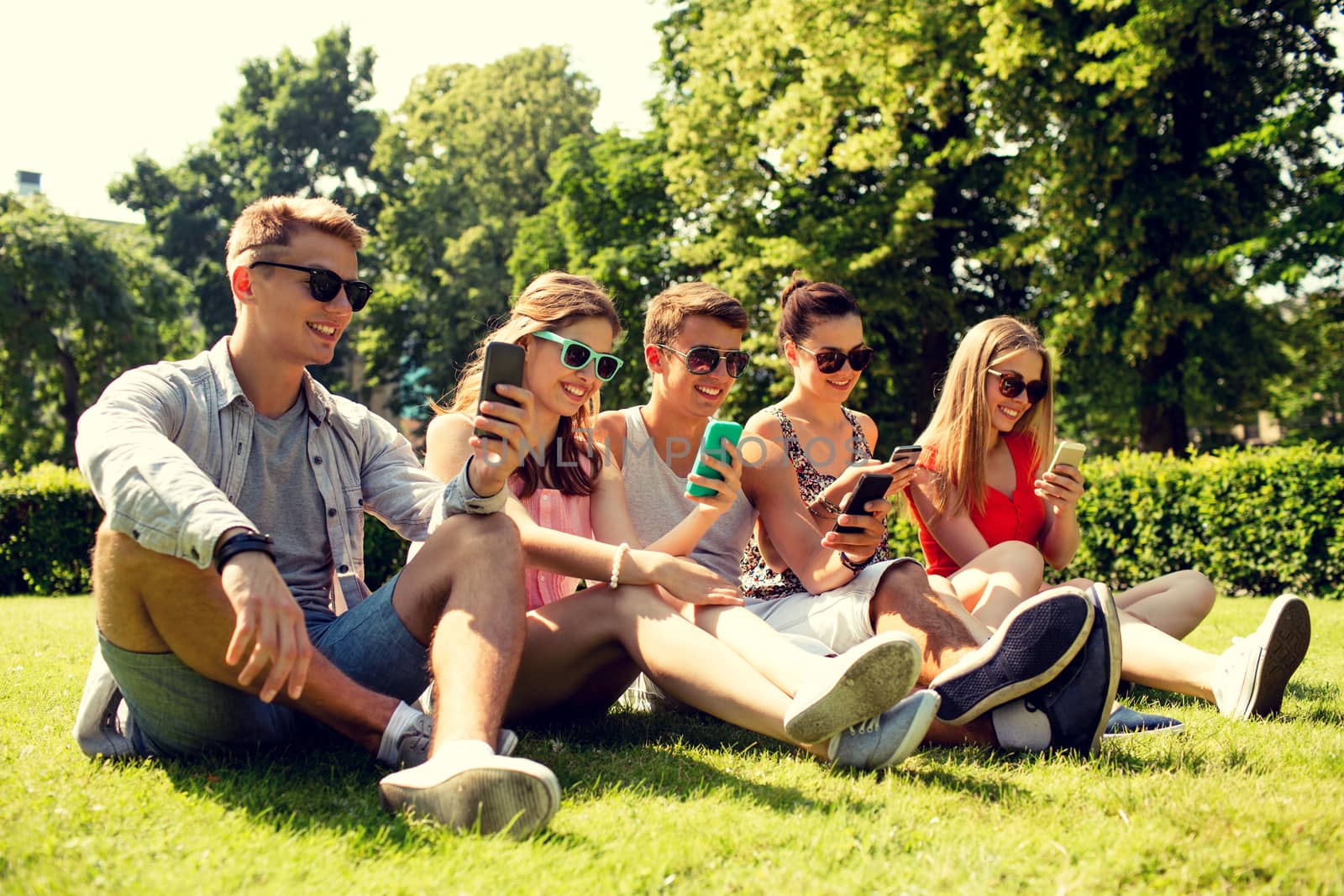 smiling friends with smartphones sitting on grass by dolgachov