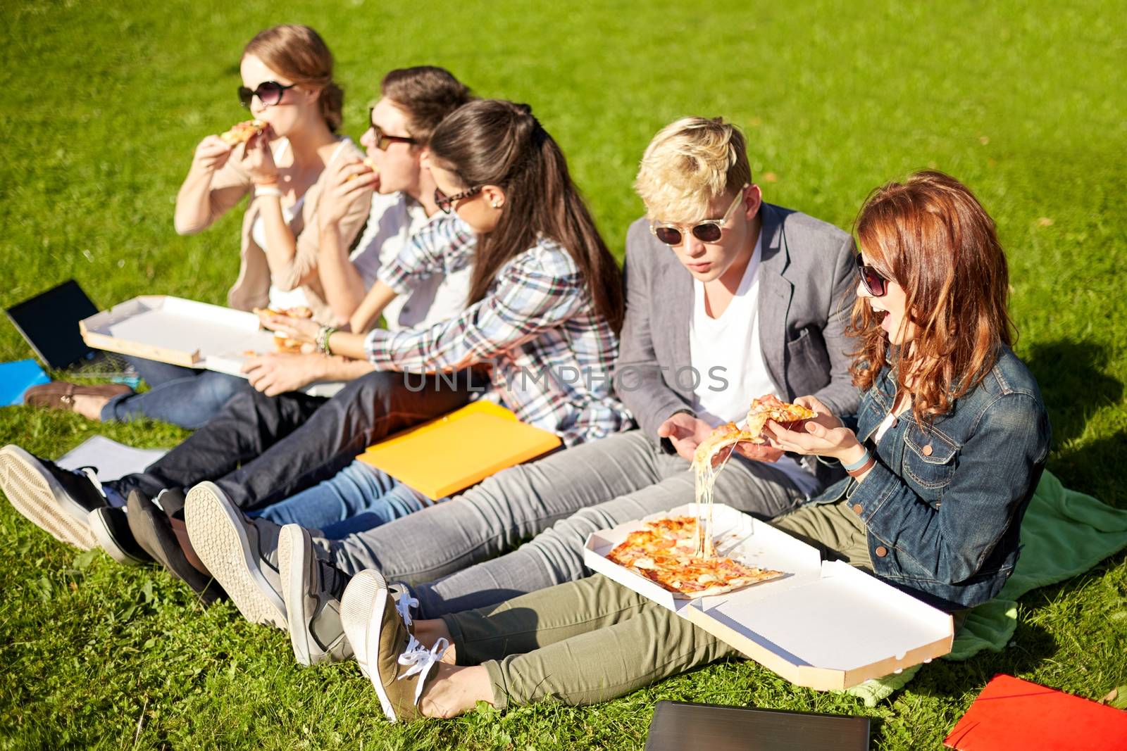 education, food, people and friendship concept - group of happy teenage students eating pizza and sitting on grass