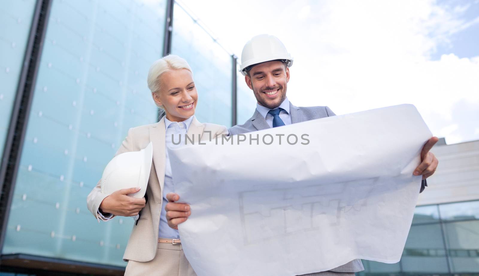 business, partnership, architecture and people concept - smiling businessman and businesswoman with blueprint and helmets on city street