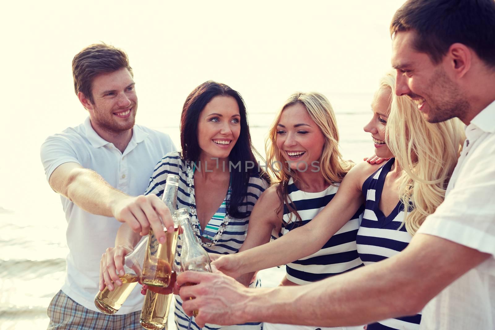 summer, holidays, tourism, drinks and people concept - group of smiling friends clinking bottles of beer or cider on beach