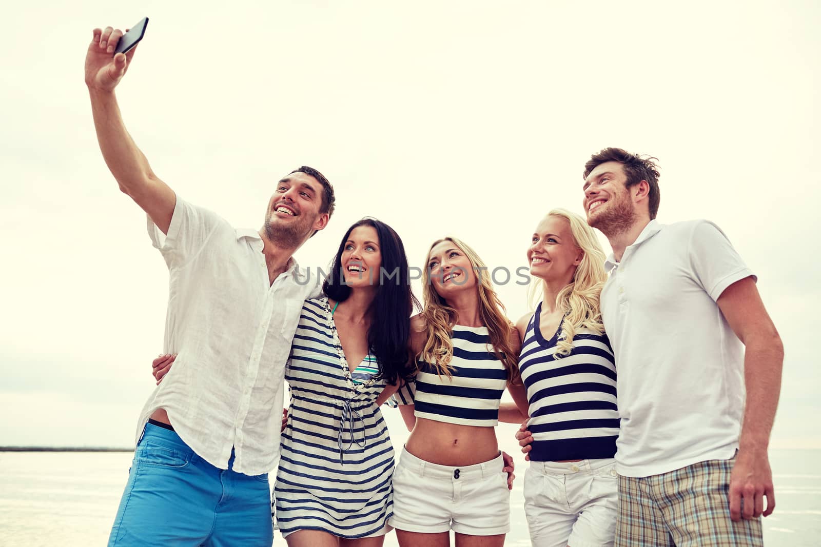 summer, sea, tourism, technology and people concept - group of smiling friends with smartphone on beach photographing and taking selfie