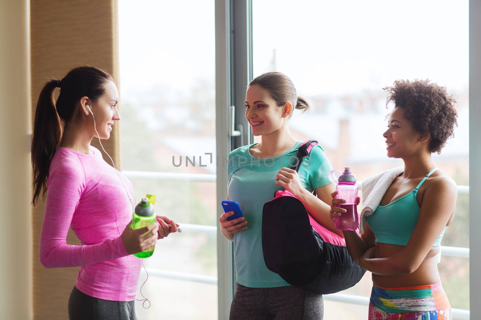 fitness, sport, training and lifestyle concept - group of happy women with bottles of water, smartphone and bag talking in gym