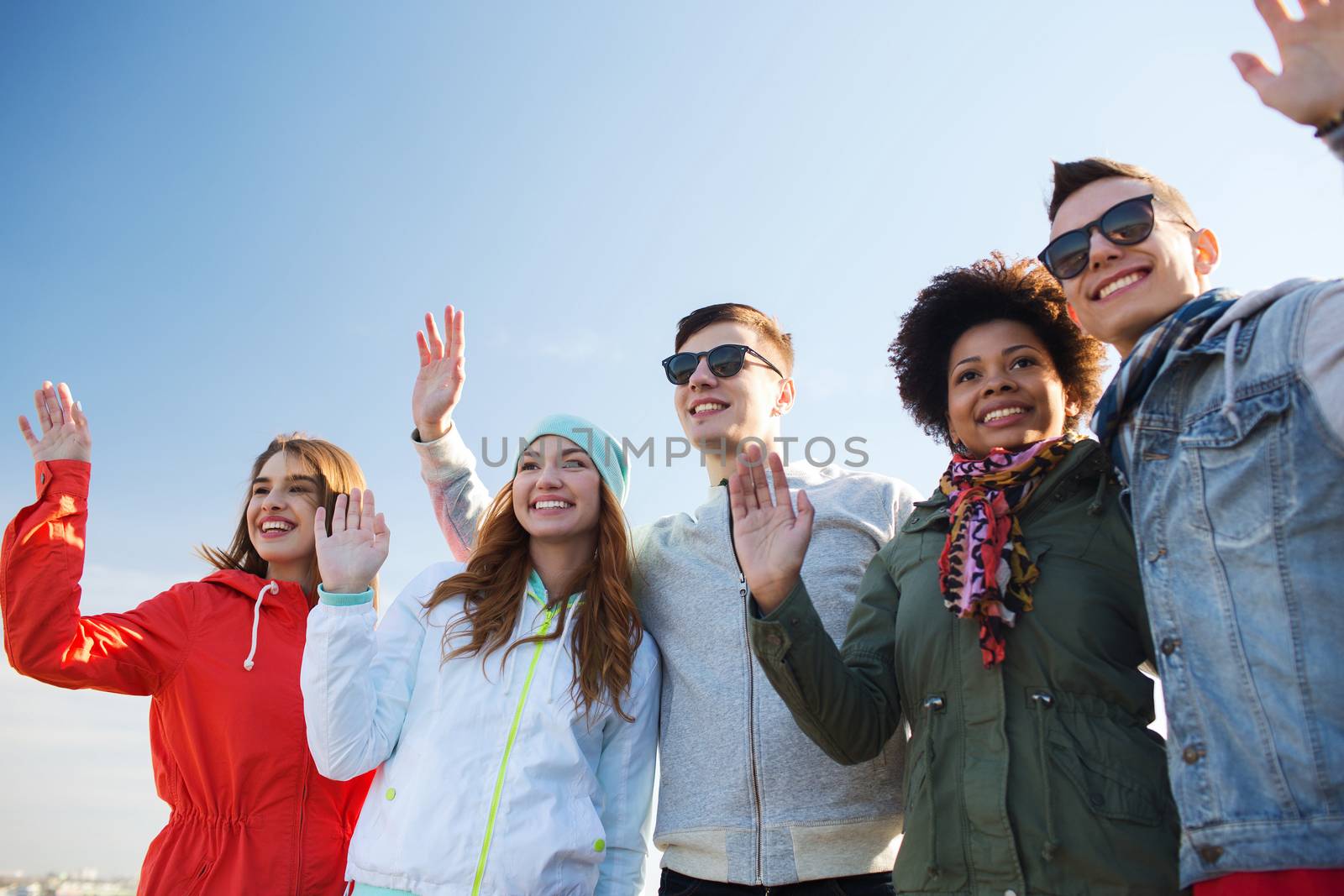 friendship, tourism, travel and people concept - group of happy teenage friends in sunglasses hugging and waving hands outdoors