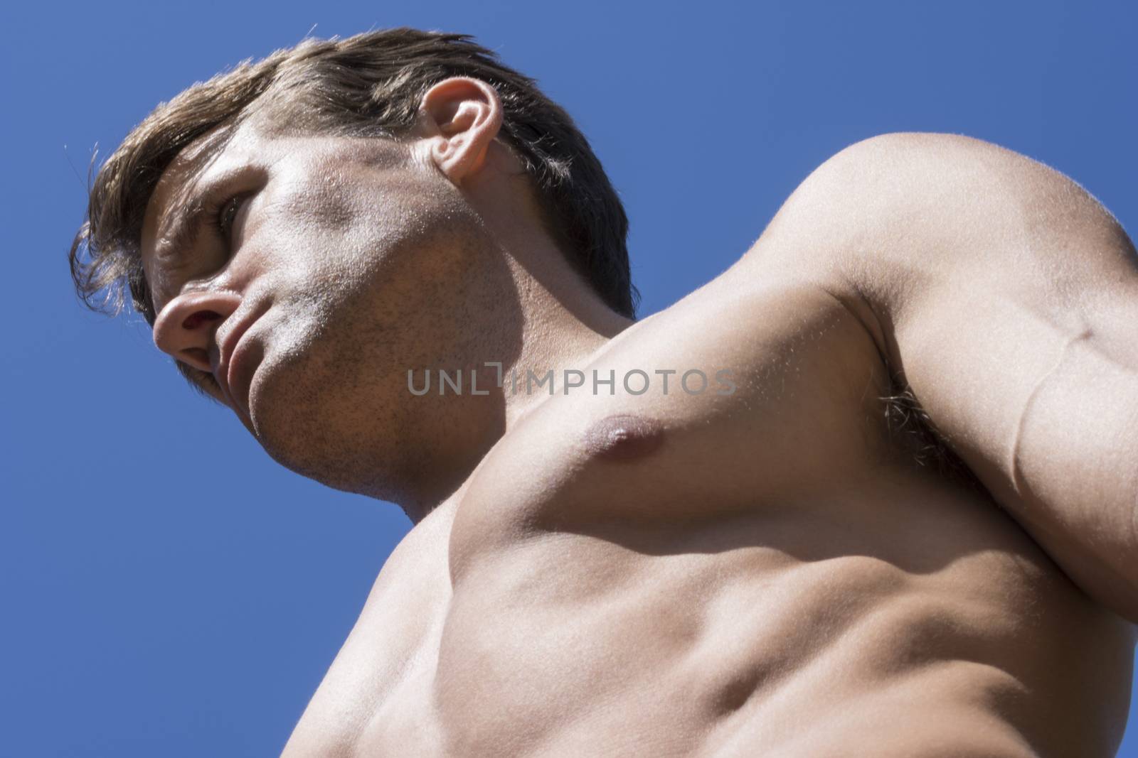 Closeup low angle with strong shadows of muscular handsome shirtless Caucasian man with bold facial expression looking forward under bright sunshine and blue sky background