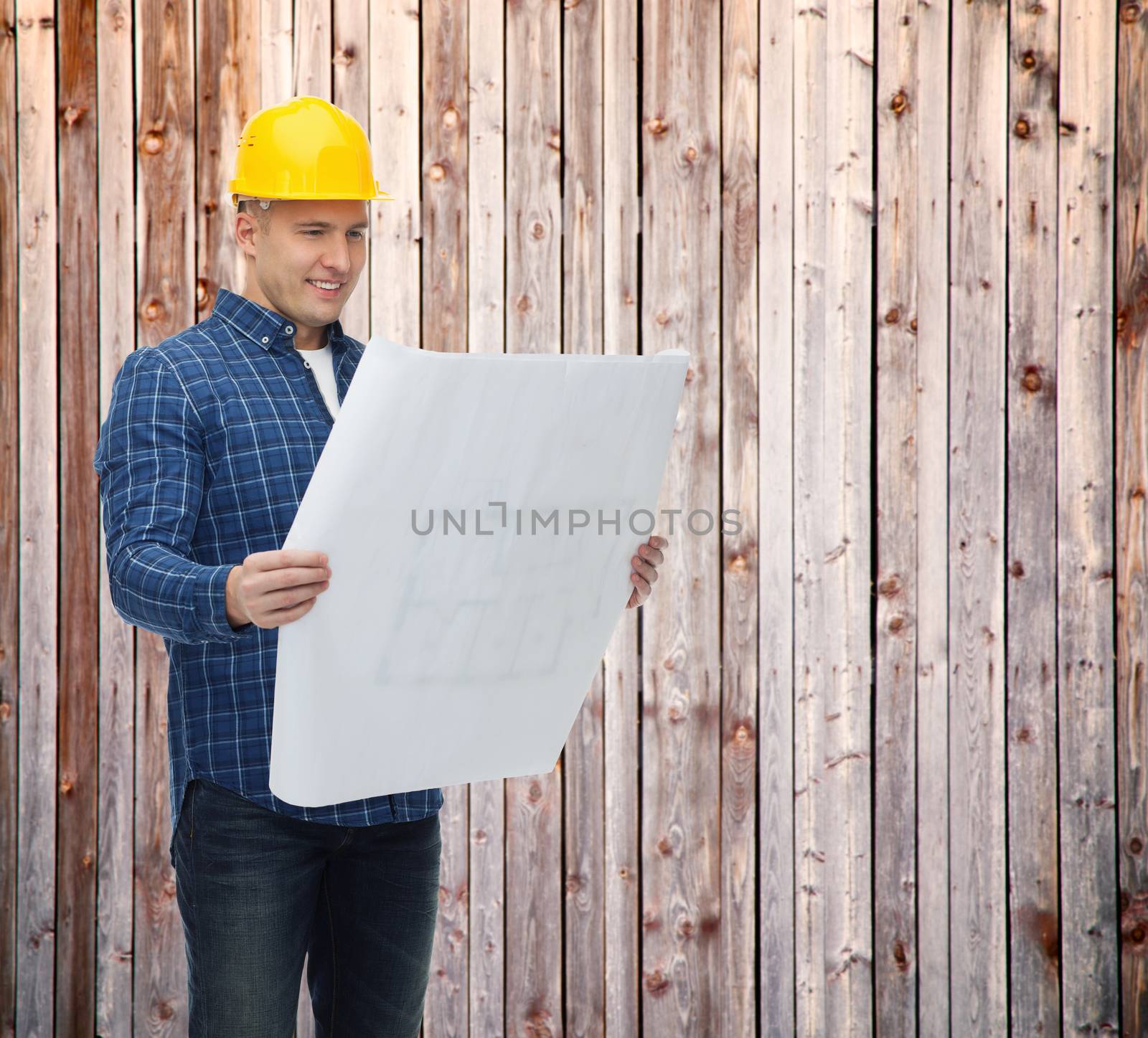 repair, construction, building, people and maintenance concept - smiling male builder or manual worker in helmet with blueprint over wooden fence background