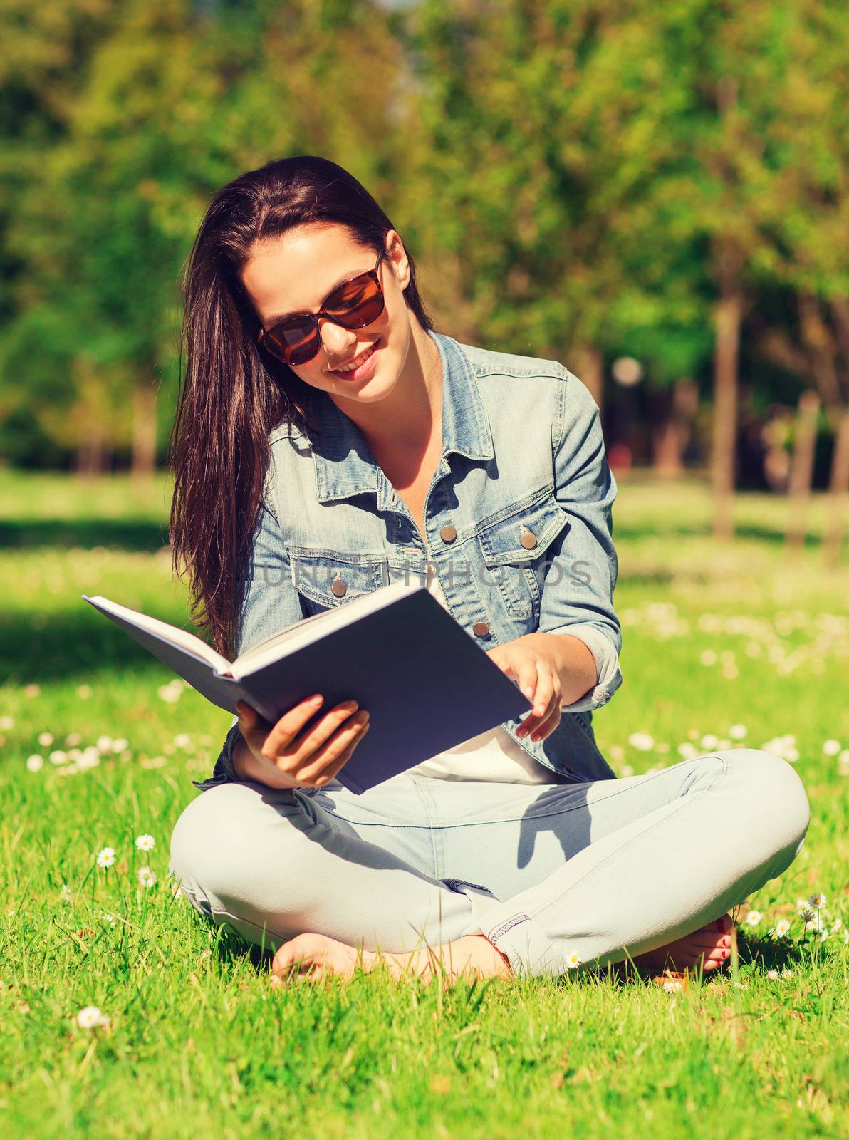 lifestyle, summer vacation, education, literature and people concept - smiling young girl reading book and sitting on grass in park