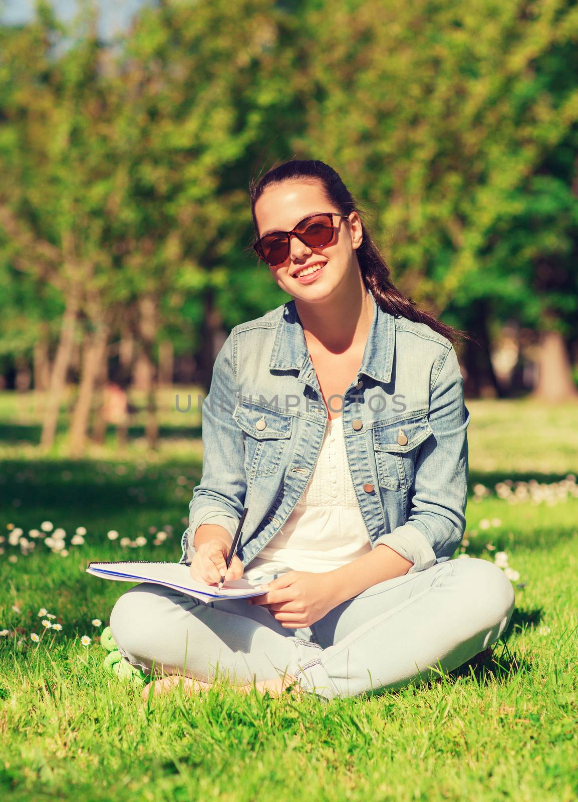 lifestyle, summer vacation, education and people concept - smiling young girl writing with pencil to notebook and sitting on grass in park