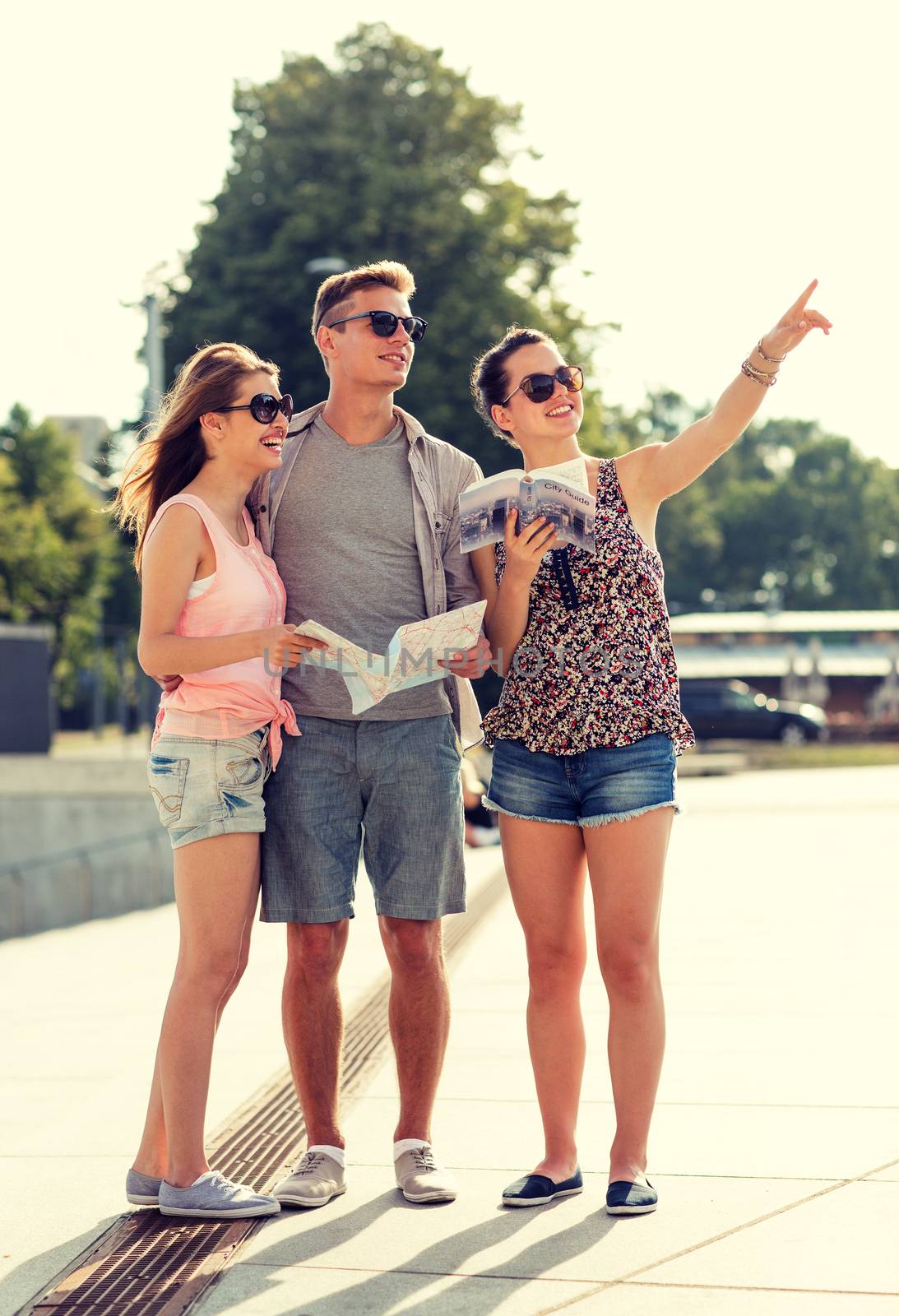 friendship, travel, tourism, vacation, summer and people concept - smiling friends with map and city guide pointing finger outdoors