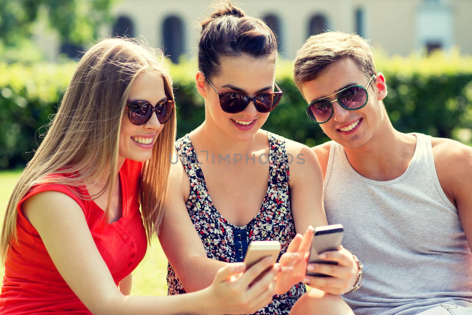 smiling friends with smartphones sitting in park by dolgachov