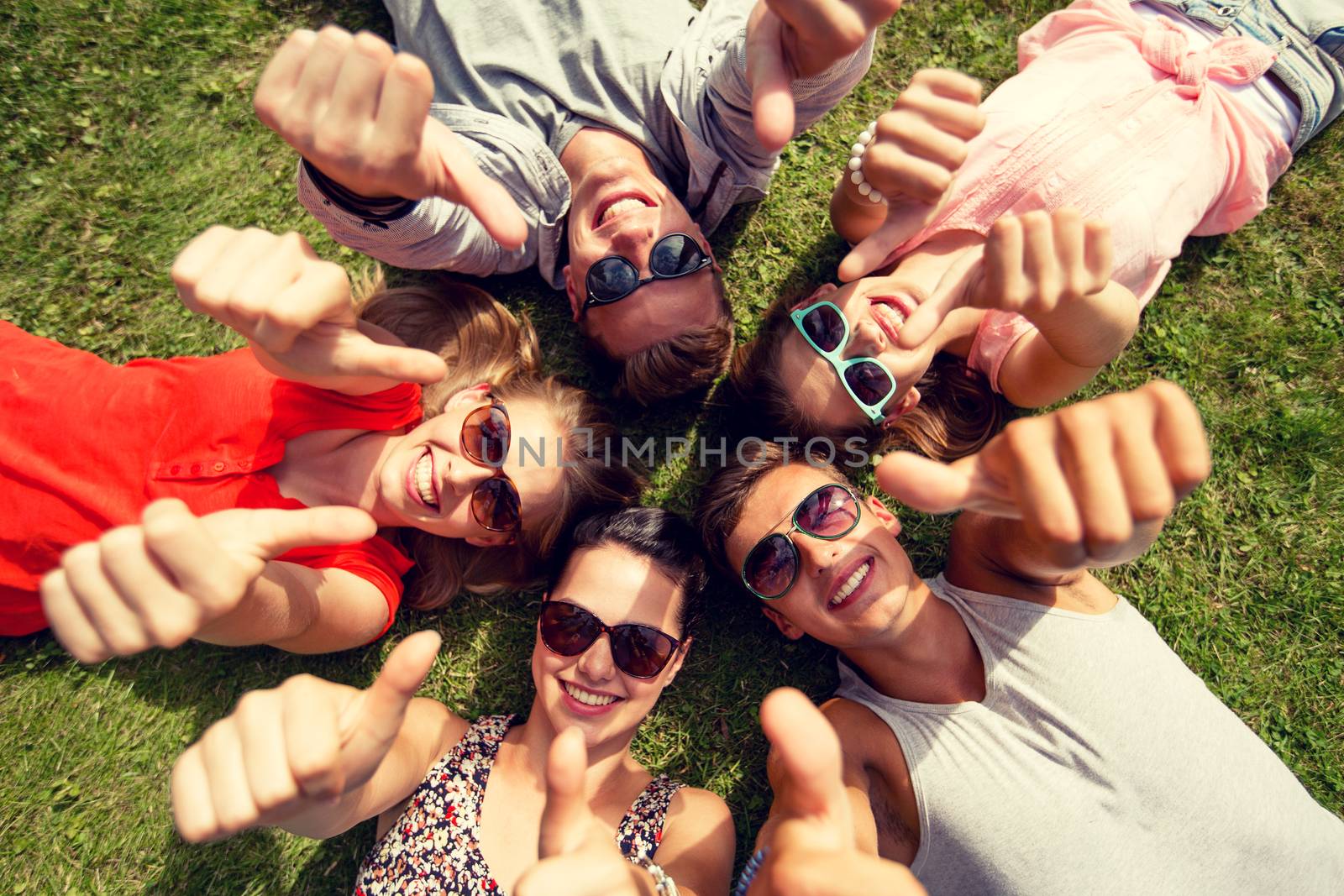 friendship, leisure, summer, gesture and people concept - group of smiling friends lying on grass in circle and showing thumbs up outdoors