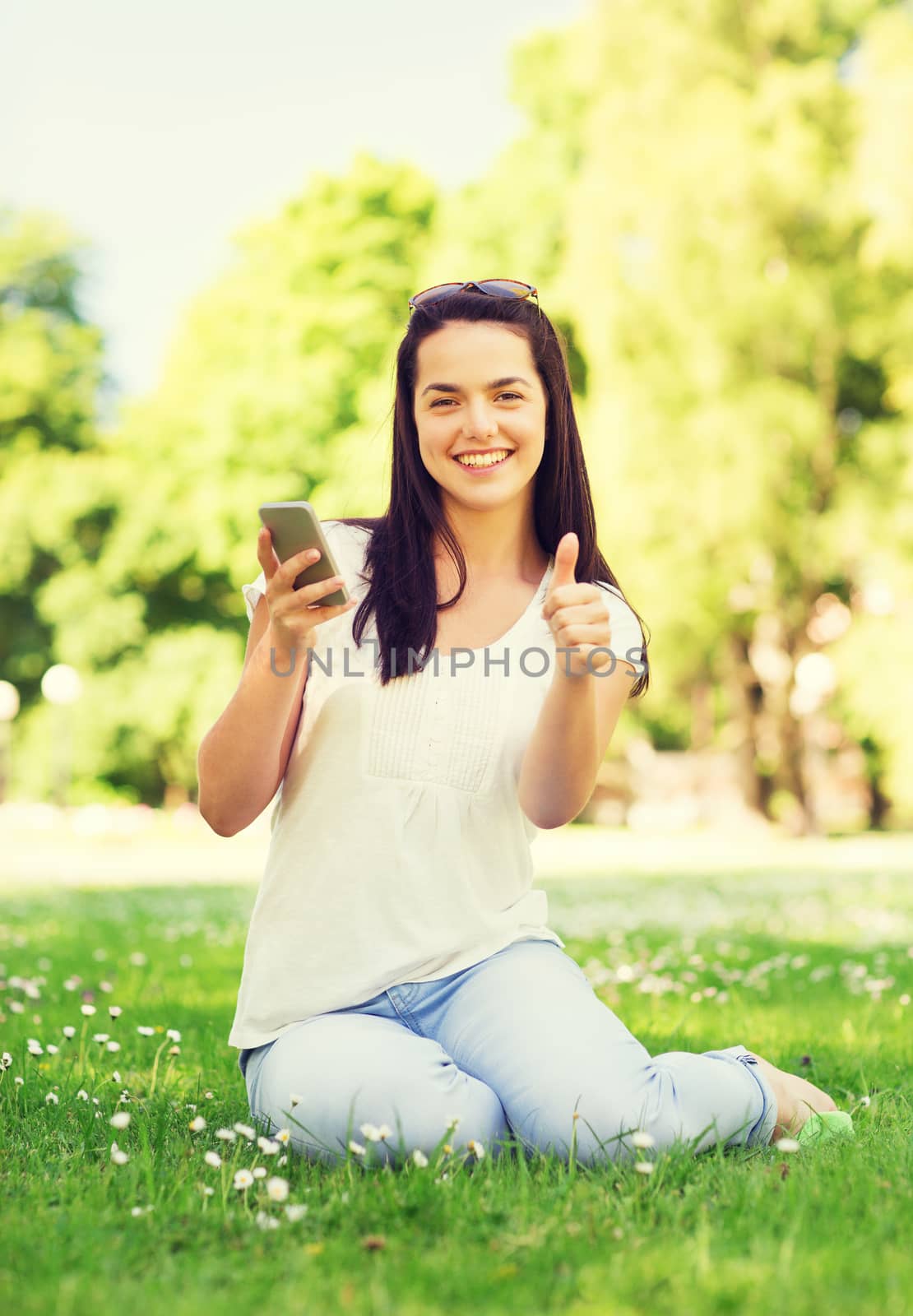 lifestyle, summer vacation, gesture, technology and people concept - smiling young girl with smartphone showing thumbs up in park