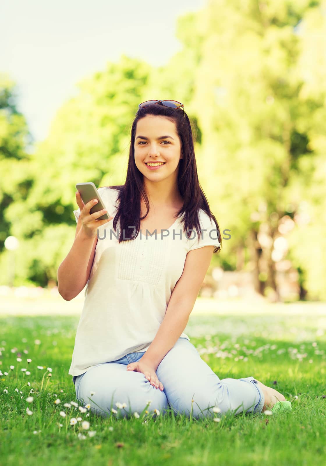 lifestyle, summer, vacation, technology and people concept - smiling young girl with smartphone sitting on grass in park