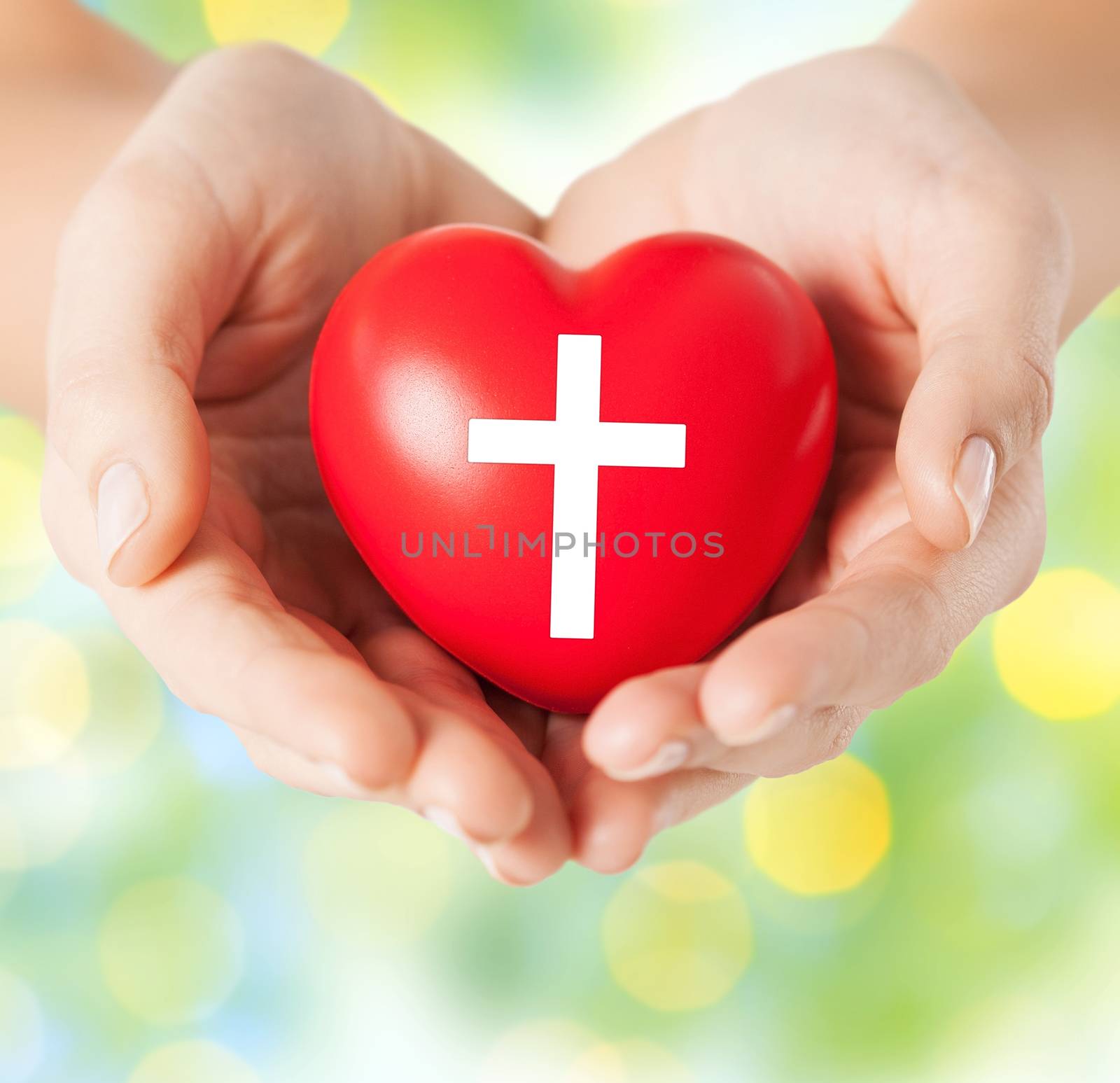 religion, christianity and charity concept - close up of female hands holding red heart with christian cross symbol over green lights background
