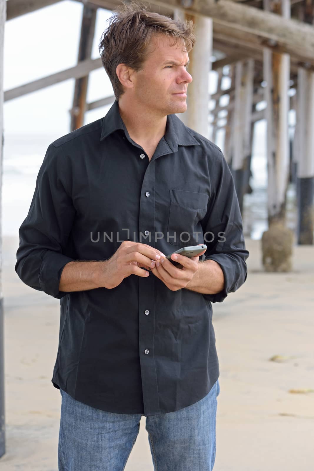 Handsome Caucasian man in casual clothing standing under pier at beach using smart phone as he waits to meet someone