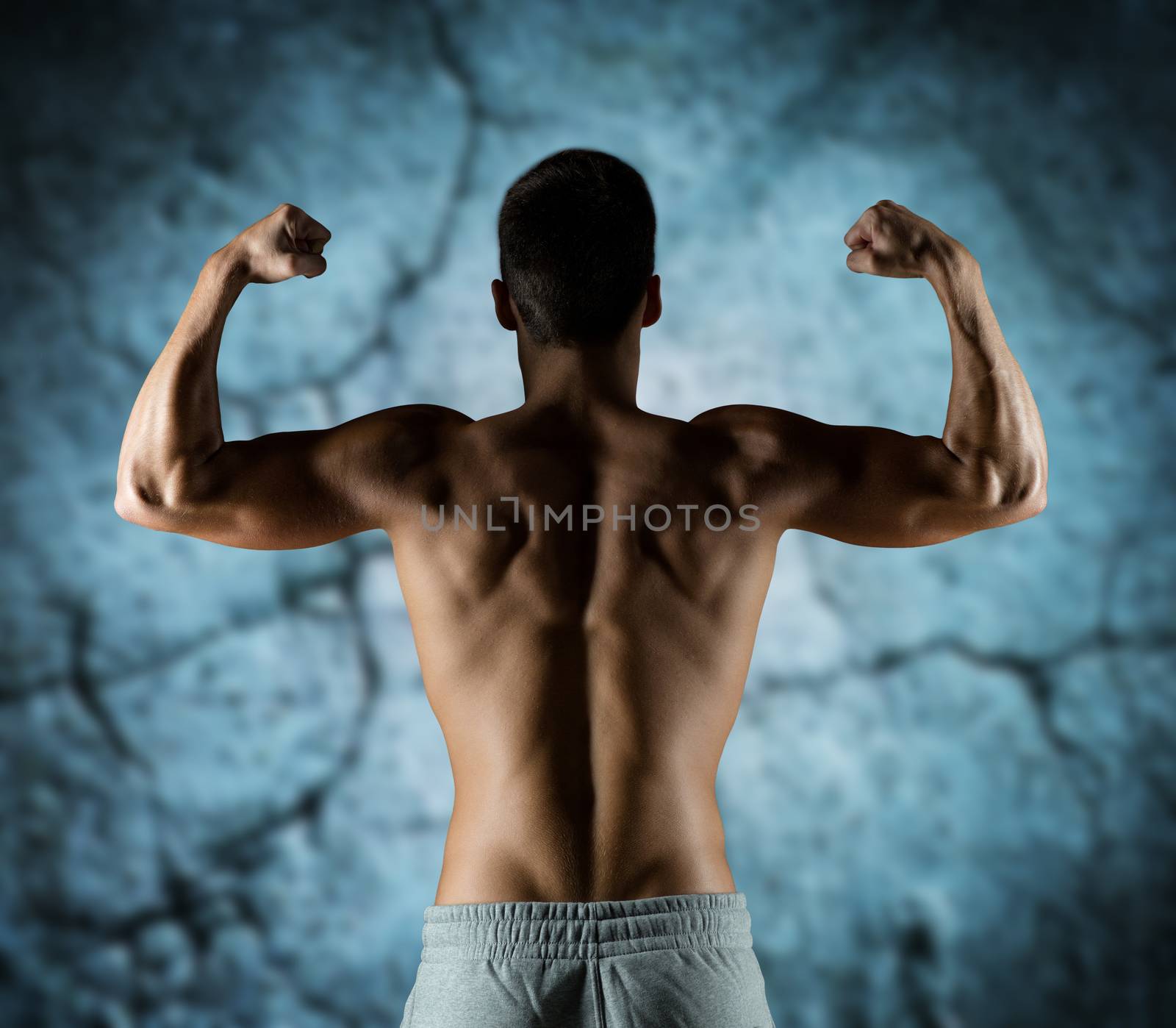 sport, fitness, bodybuilding, strength and people concept - young man or bodybuilder showing biceps over concrete wall background from back