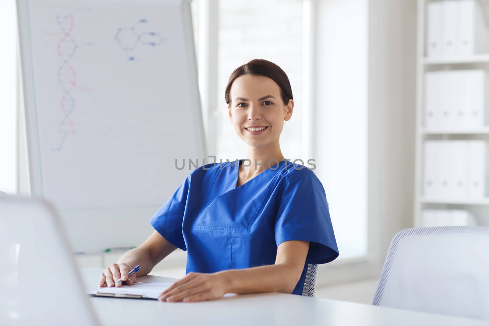 medicine, people and healthcare concept - happy female doctor or nurse writing medical report to clipboard at hospital