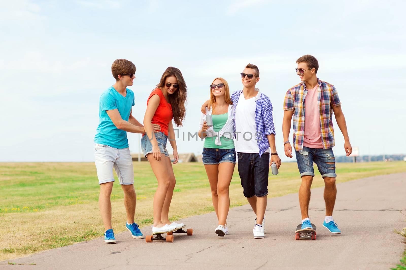 holidays, vacation, love and friendship concept - group of smiling teenagers walking and riding on skateboards outdoors