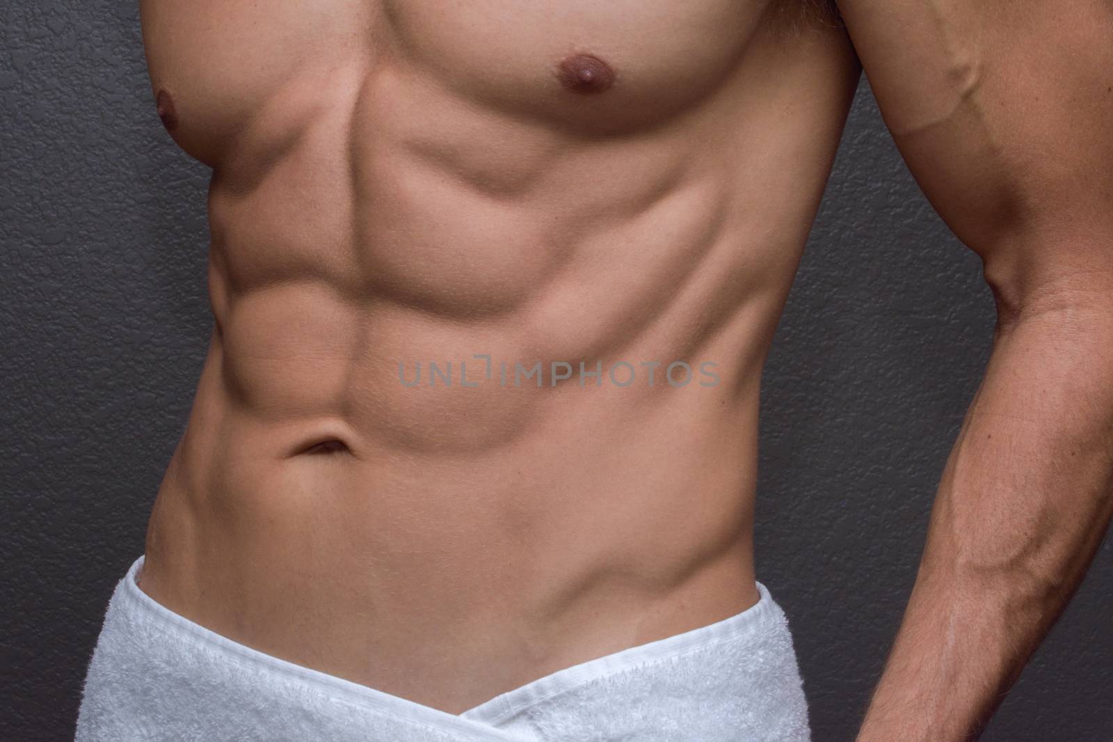 Closeup of sexy torso showing muscular lean abdominals and chest of tan Caucasian man with white towel wrapped around waist next to textured gray wall background