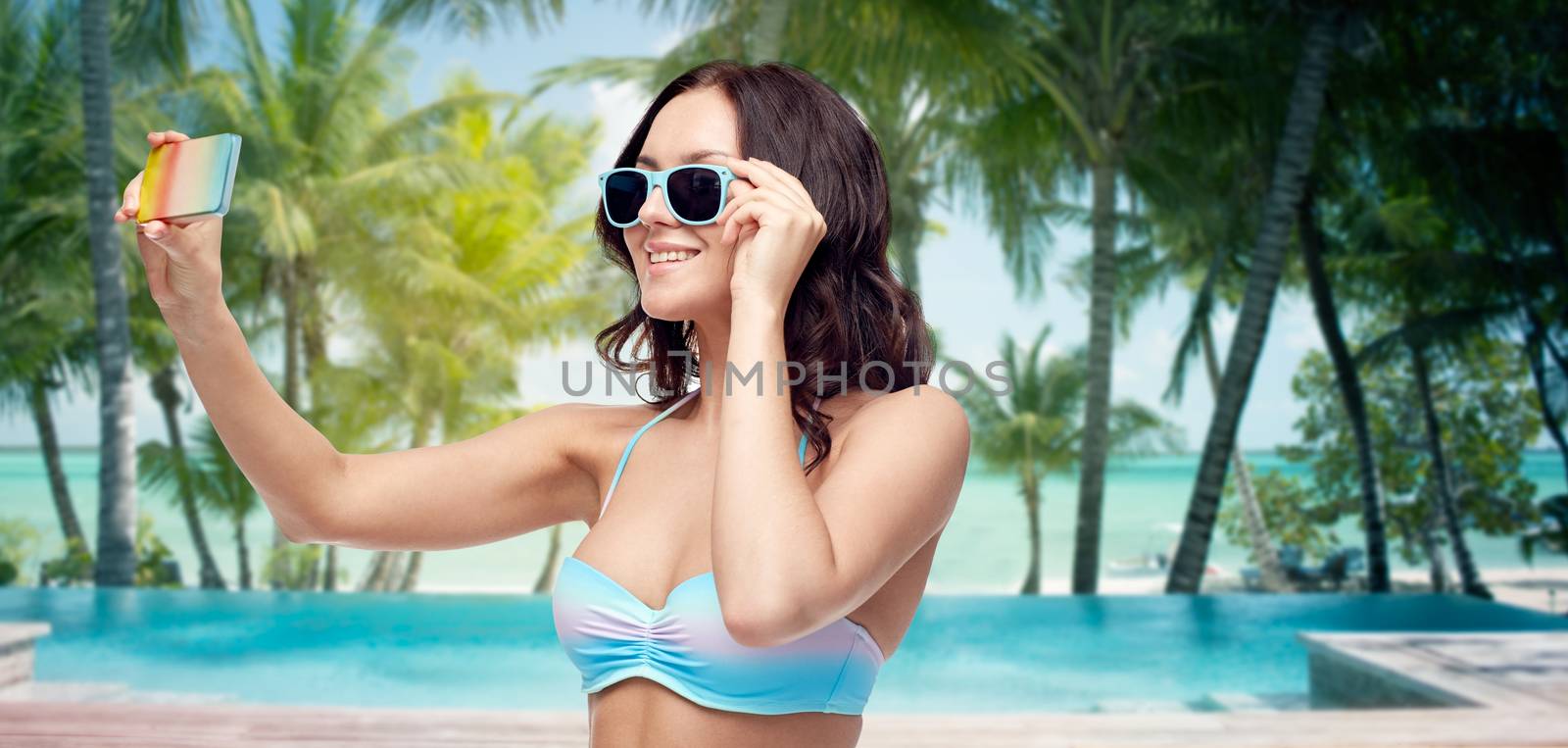 people, technology, travel, tourism and summer concept - happy young woman in bikini swimsuit and sunglasses taking selfie with smatphone over swimming pool on beach with palm trees background