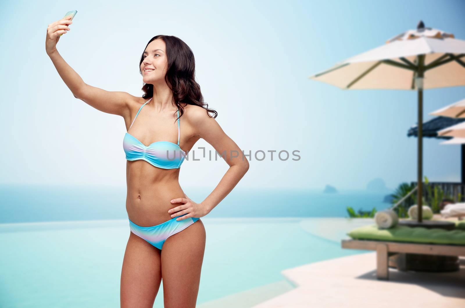 people, technology, travel, tourism and summer concept - happy young woman in bikini swimsuit taking selfie with smatphone over infinity pool with parasol and sunbed at sea side background