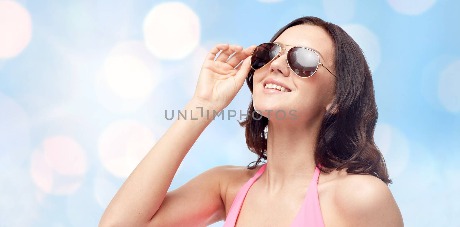 people, fashion, swimwear, summer and beach concept - happy young woman in sunglasses and pink swimsuit looking up over blue holidays lights background