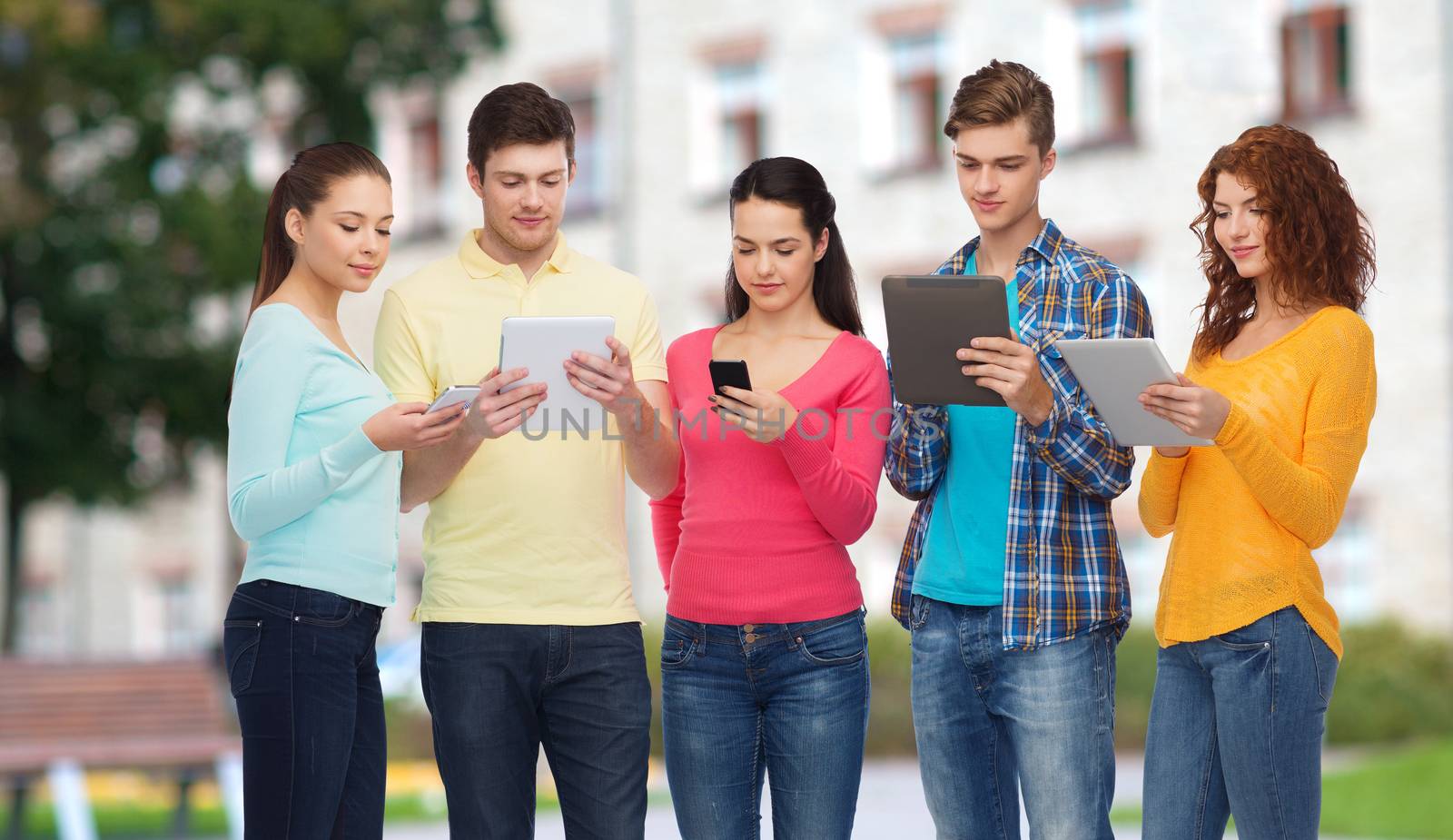 friendship, technology, education, school and people concept - group of smiling teenagers with smartphones and tablet pc computers over campus background