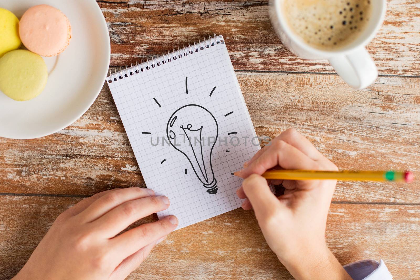 business, education and people concept - close up of female hands with pencil, coffee and cookies drawing lighting bulb to notebook on table