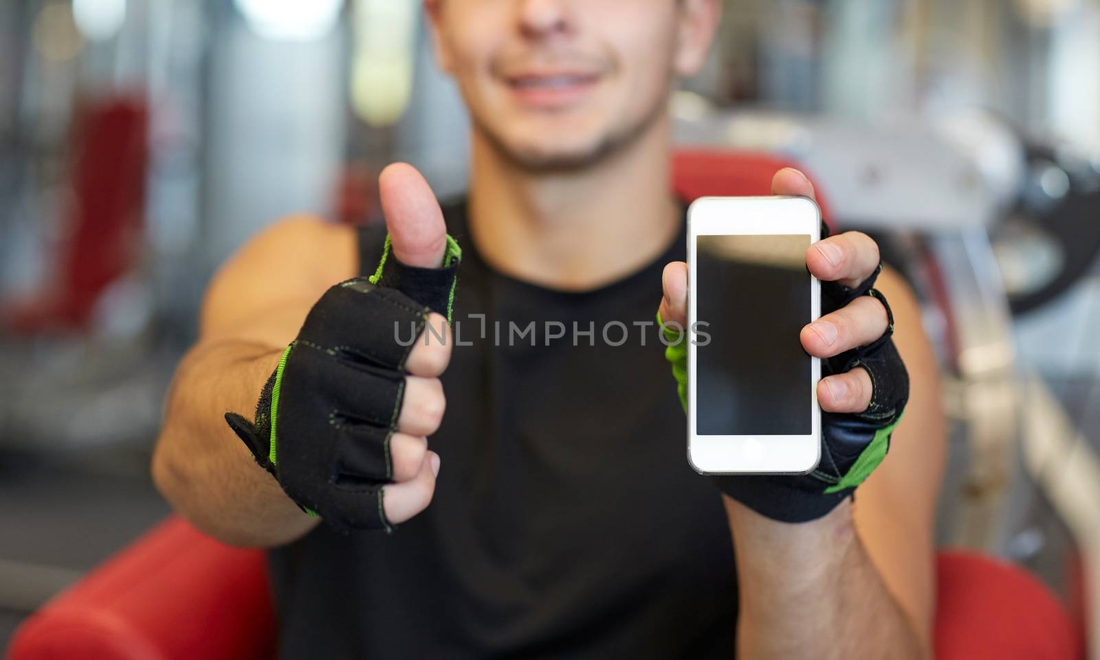 young man with smartphone showing thumbs up in gym by dolgachov