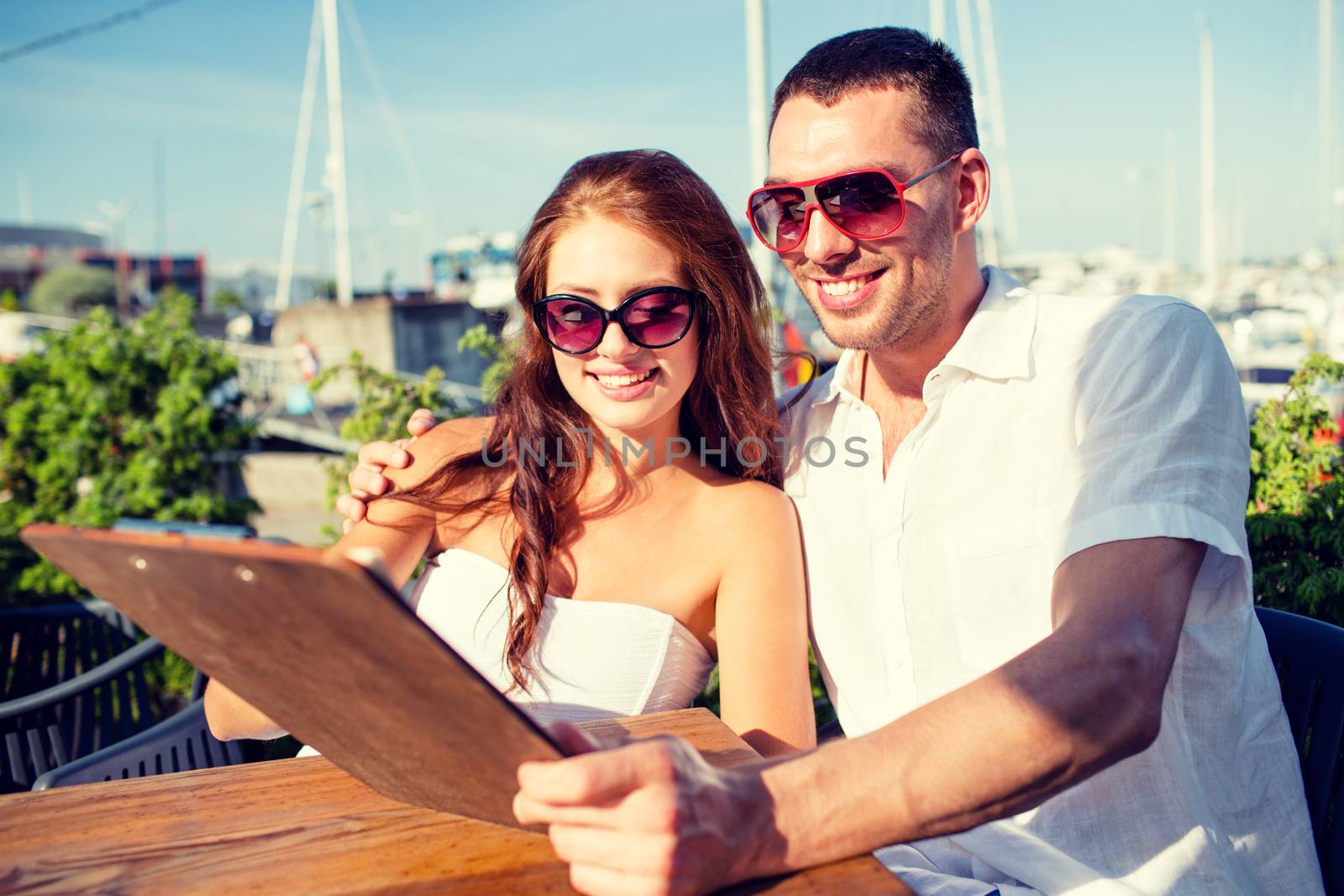 love, dating, people and food concept - smiling couple wearing sunglasses looking at menu on cafe terrace