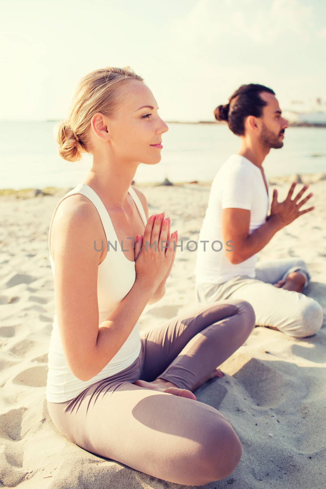 fitness, sport, friendship and lifestyle concept - smiling couple making yoga exercises sitting on sand outdoors