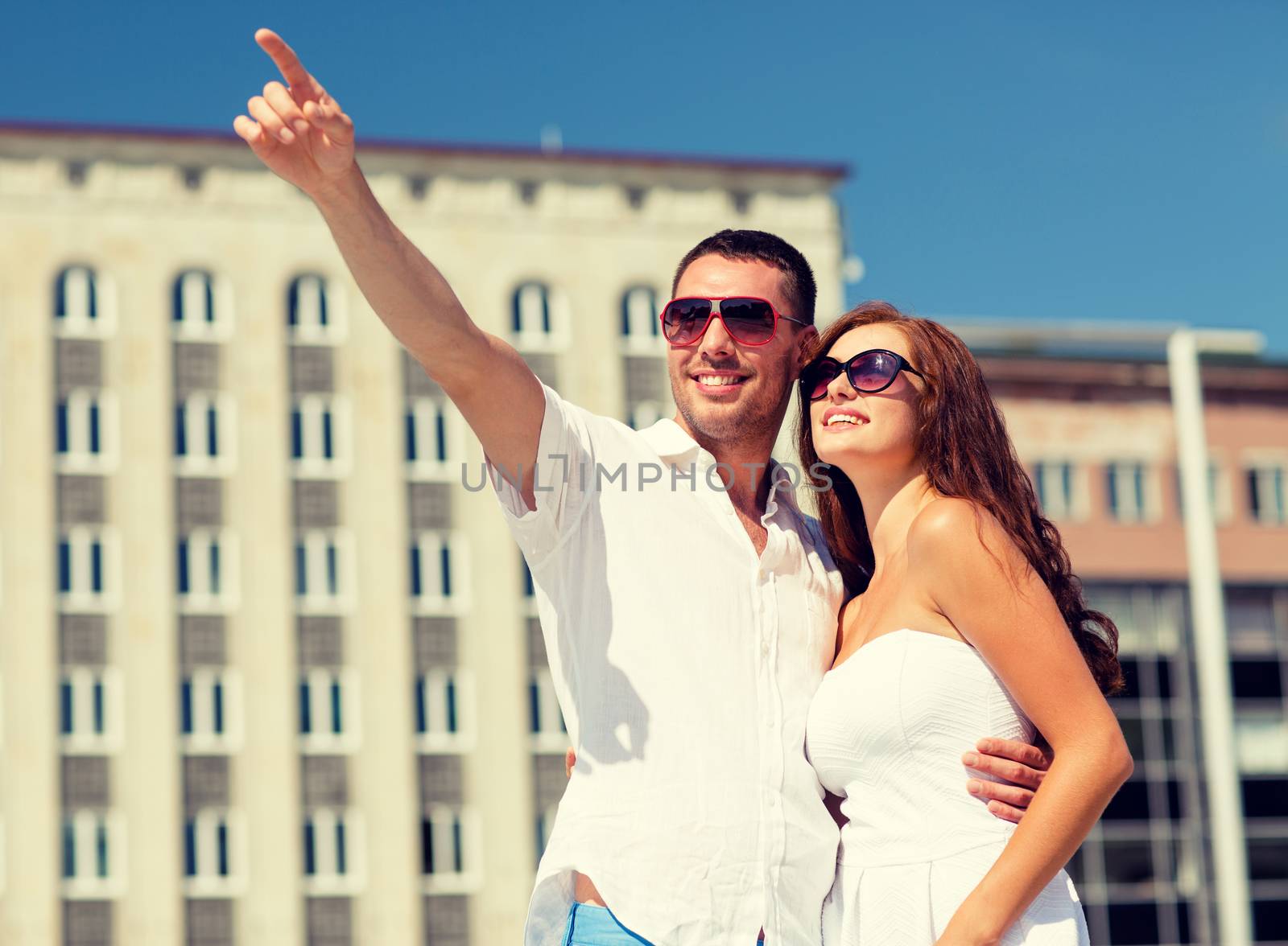 love, travel, tourism, people and friendship concept - smiling couple wearing sunglasses hugging and pointing finger in city