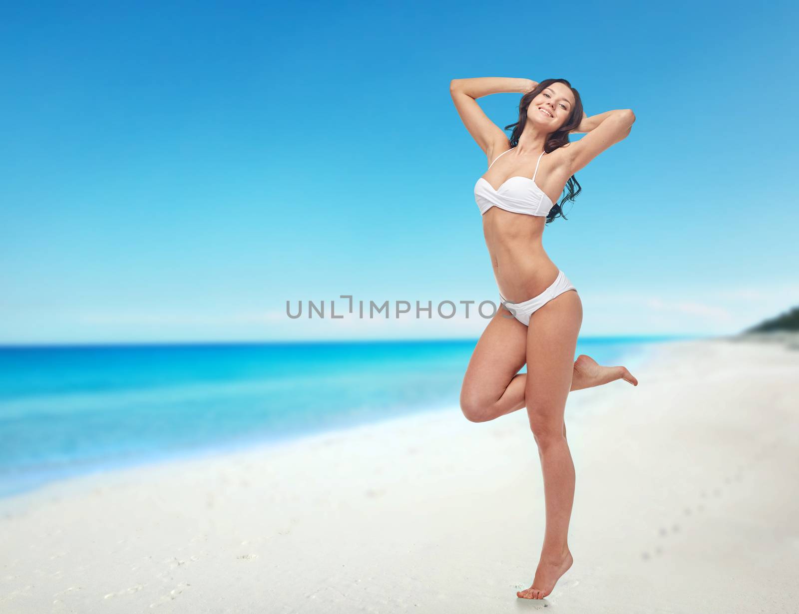 people, swimwear, tourism, travel and summer concept - happy young woman posing in white bikini swimsuit with raised hands and standing on one leg over beach background