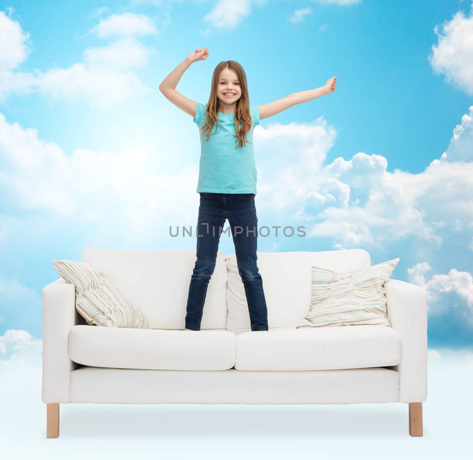 home, leisure, people and happiness concept - smiling little girl jumping and dancing on sofa over blue sky and white clouds background