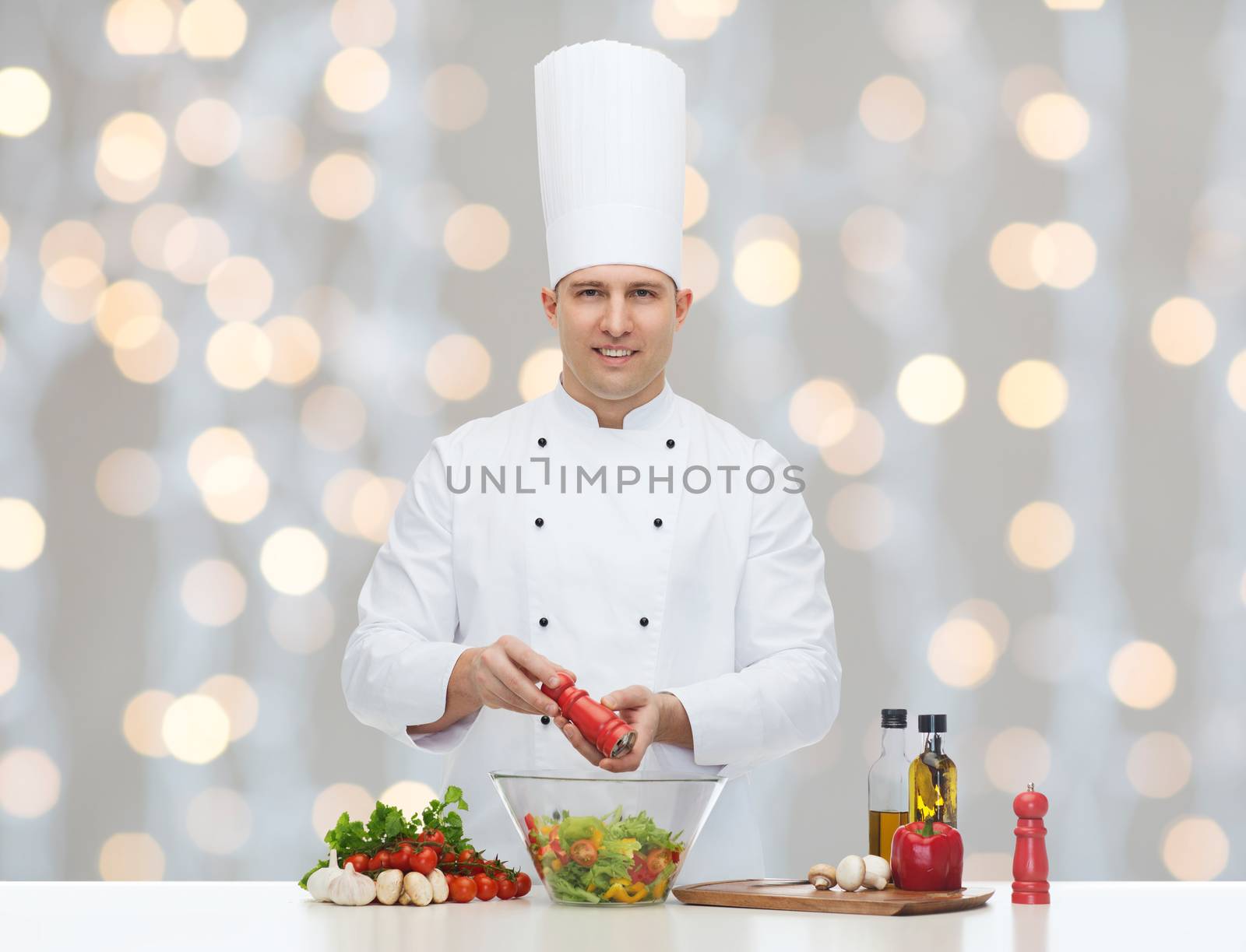 profession, vegetarian, food and people concept - happy male chef cooking and seasoning vegetable salad over christmas holidays lights background