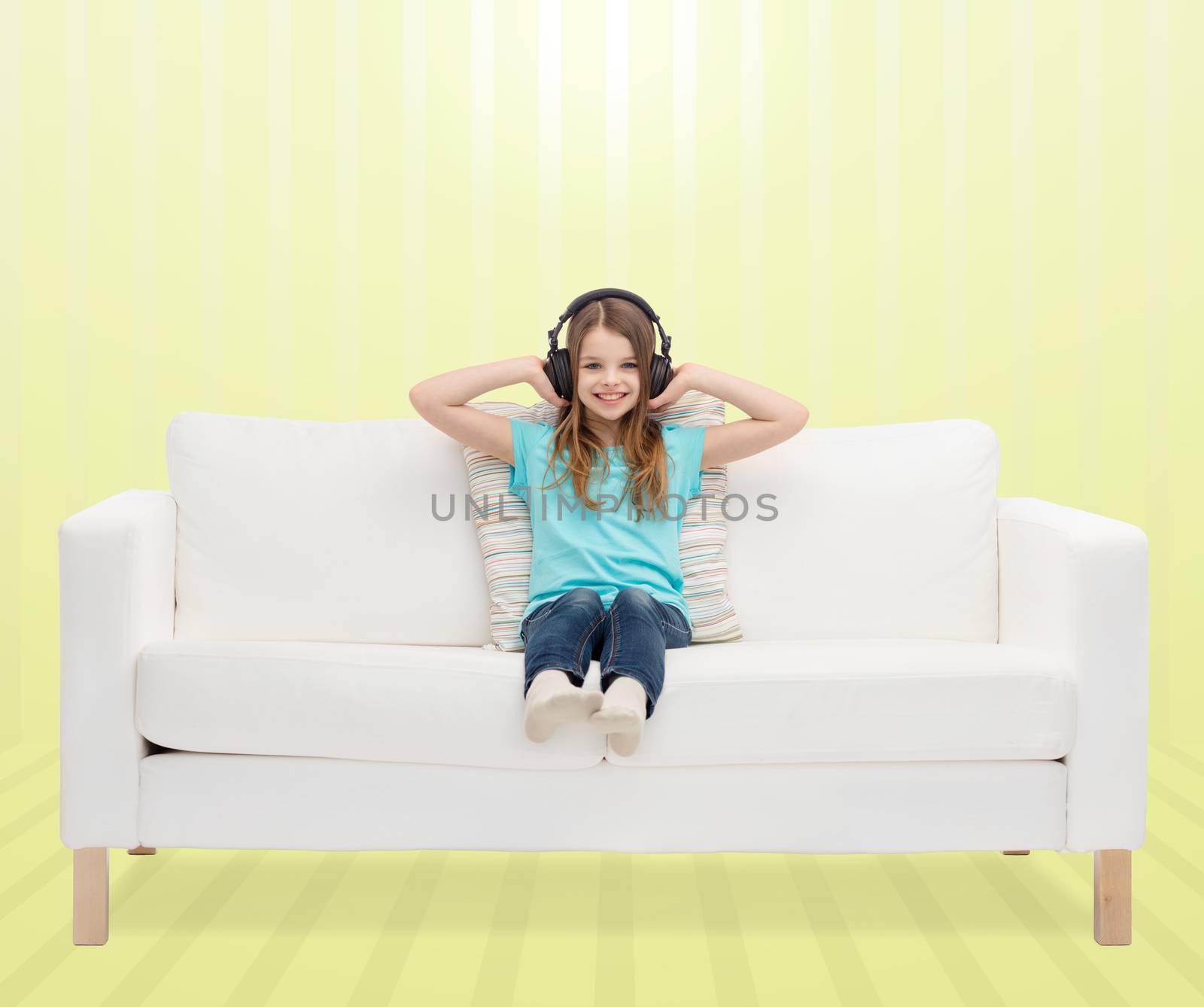 leisure, technology, music and childhood concept - smiling little girl in headphones listening to music sitting on sofa over yellow striped background