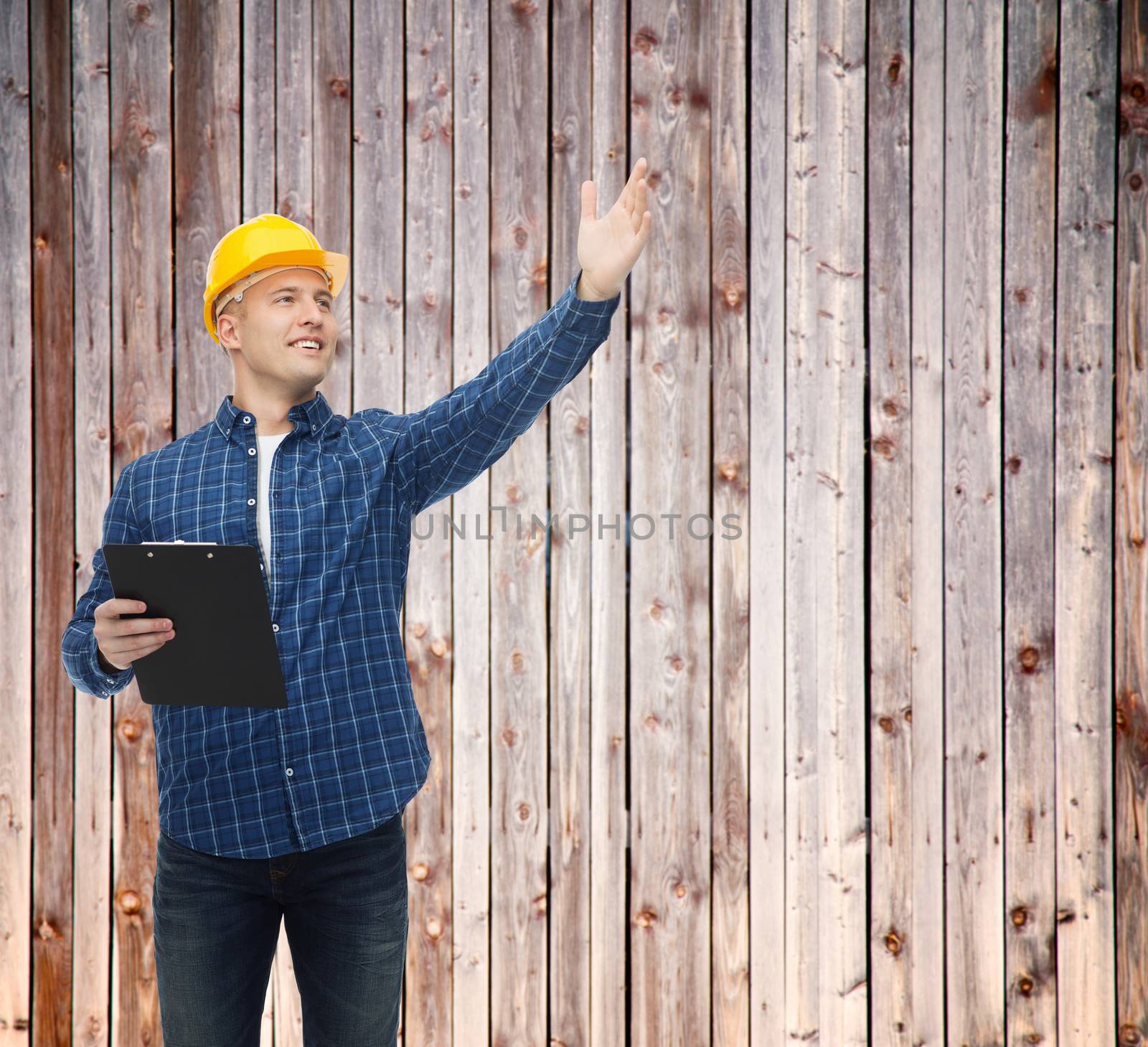 repair, construction, building, people and maintenance concept - smiling male builder or manual worker in helmet with clipboard pointing hand over wooden fence background