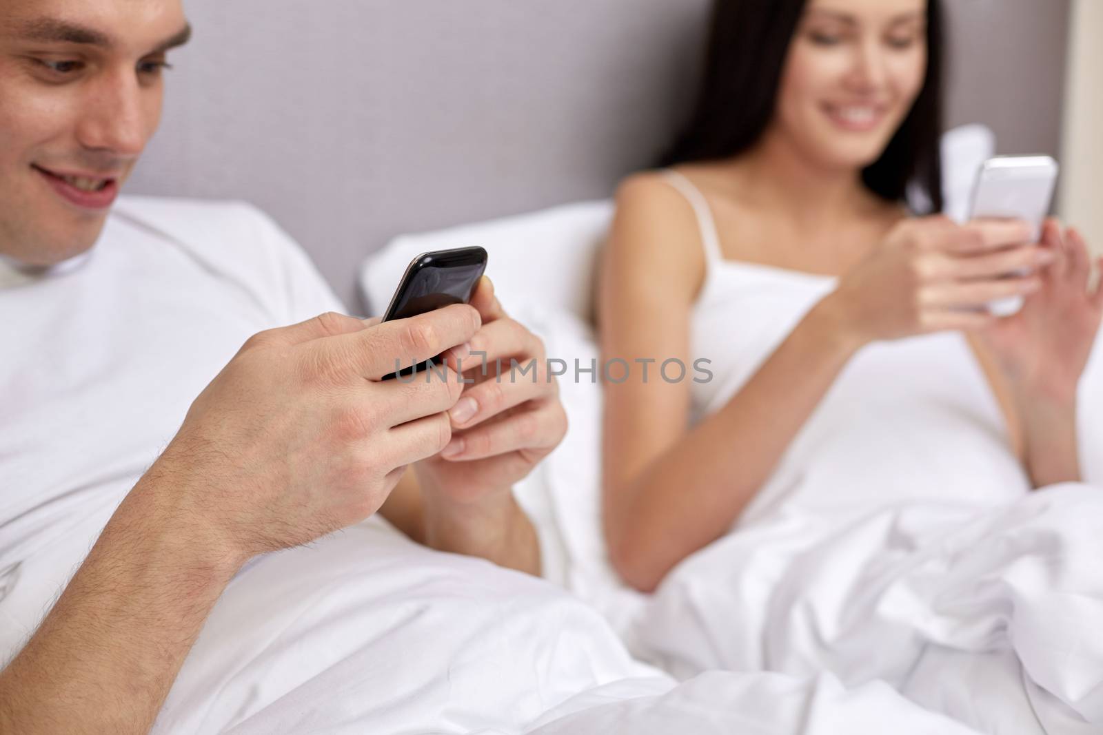 people, bedtime, technology and relations concept - close up of smiling couple in bed with smartphones texting message