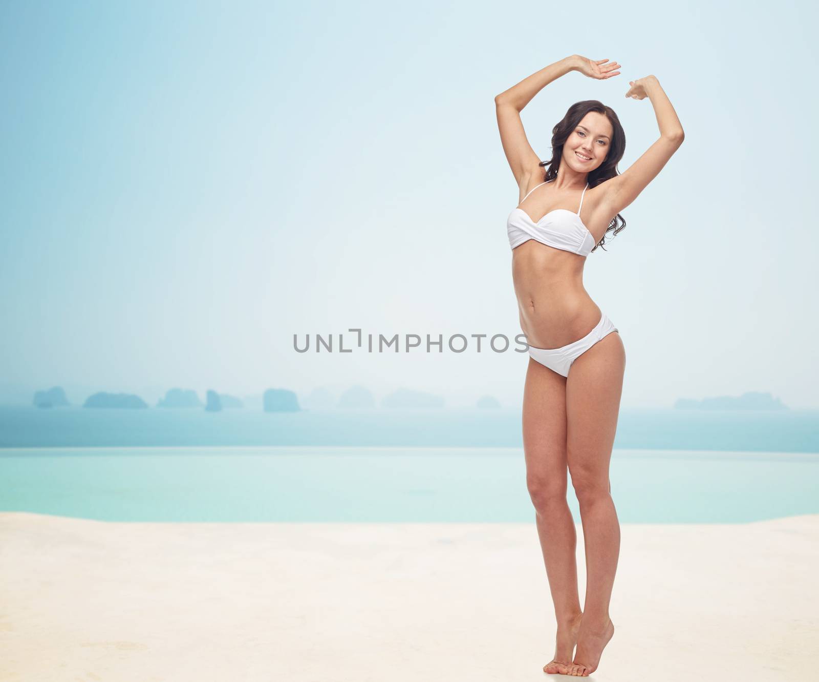 people, fashion, swimwear, summer and beach concept - happy young woman posing in white bikini swimsuit with raised hands over infinity pool at beach resort